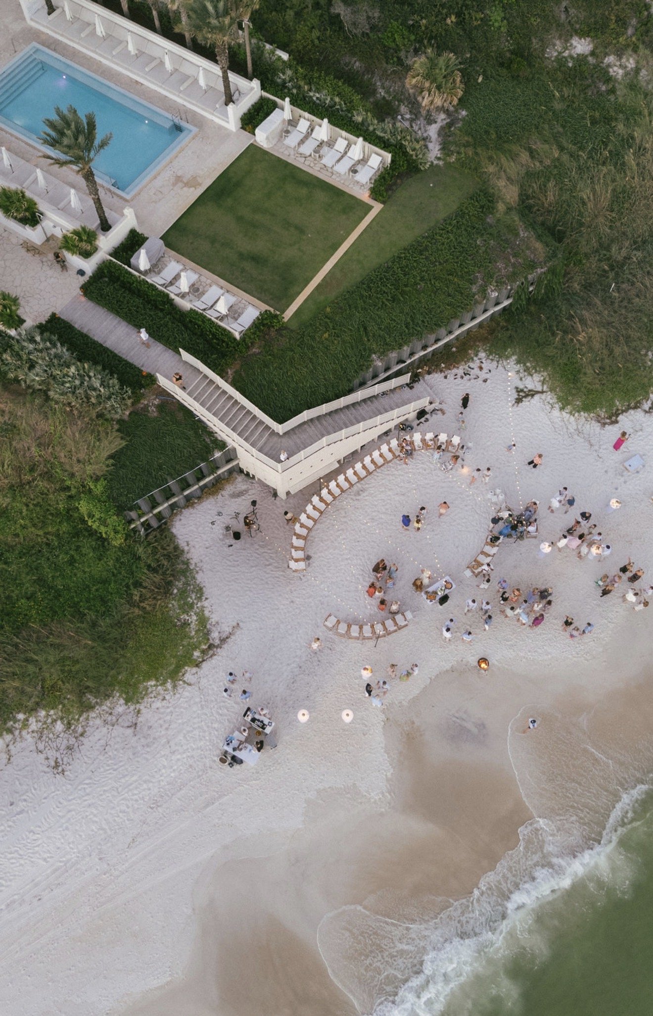 Aerial view of a beach with white sand, dotted with beach chairs and umbrellas, and a shoreline with waves crashing onto the sand. There is a deck with more chairs and umbrellas, leading to a green grassy area, and a swimming pool with lounge chairs 