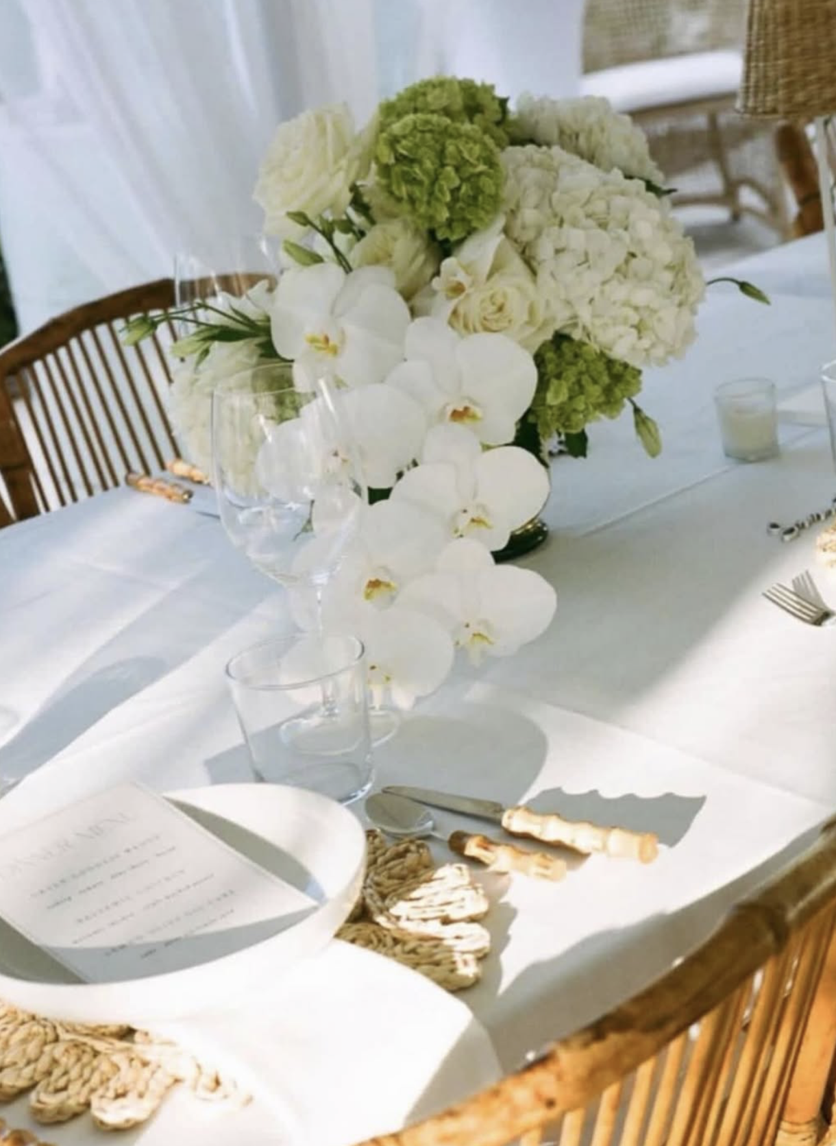 A table decorated with a large floral centerpiece featuring white roses, orchids, and hydrangeas, set with glassware, cutlery, and a menu, in a bright and elegant setting.