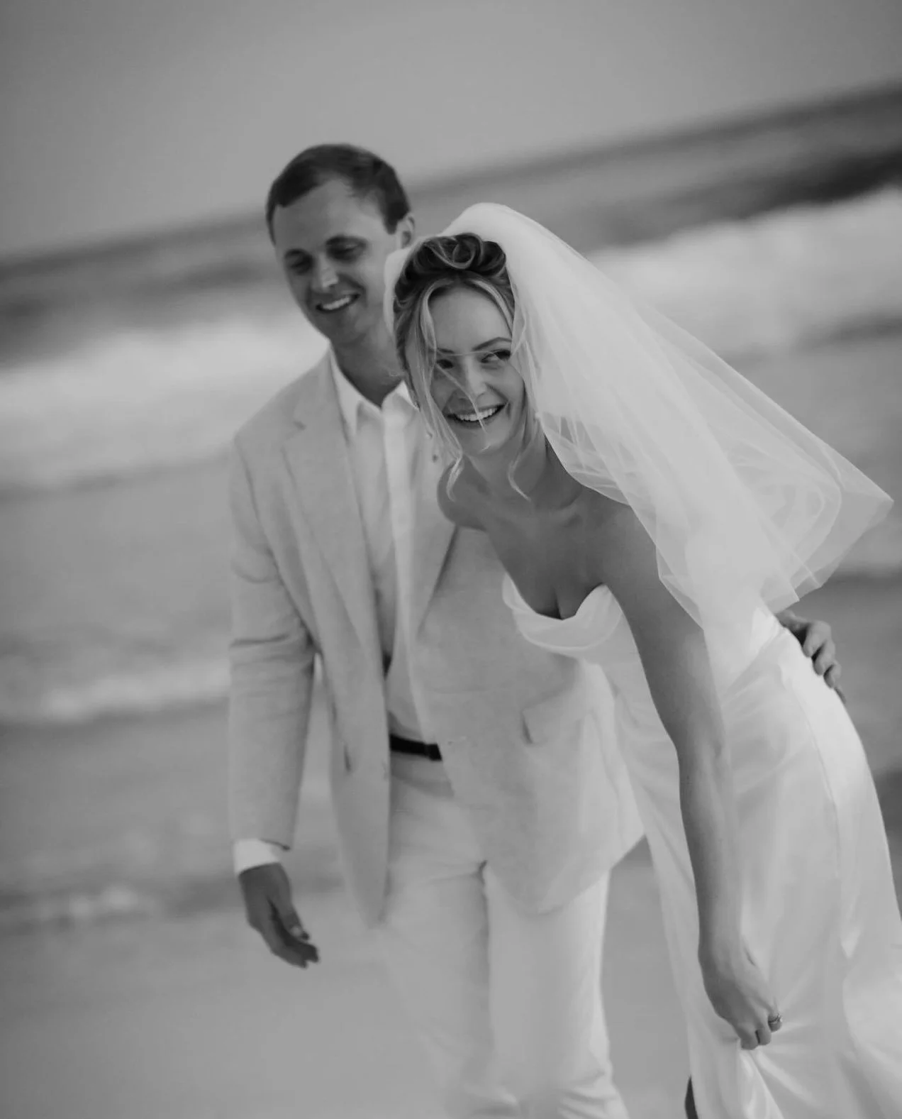 A black-and-white photo of a smiling bride and groom on the beach, with the bride leaning forward and holding her dress as they enjoy their wedding day.