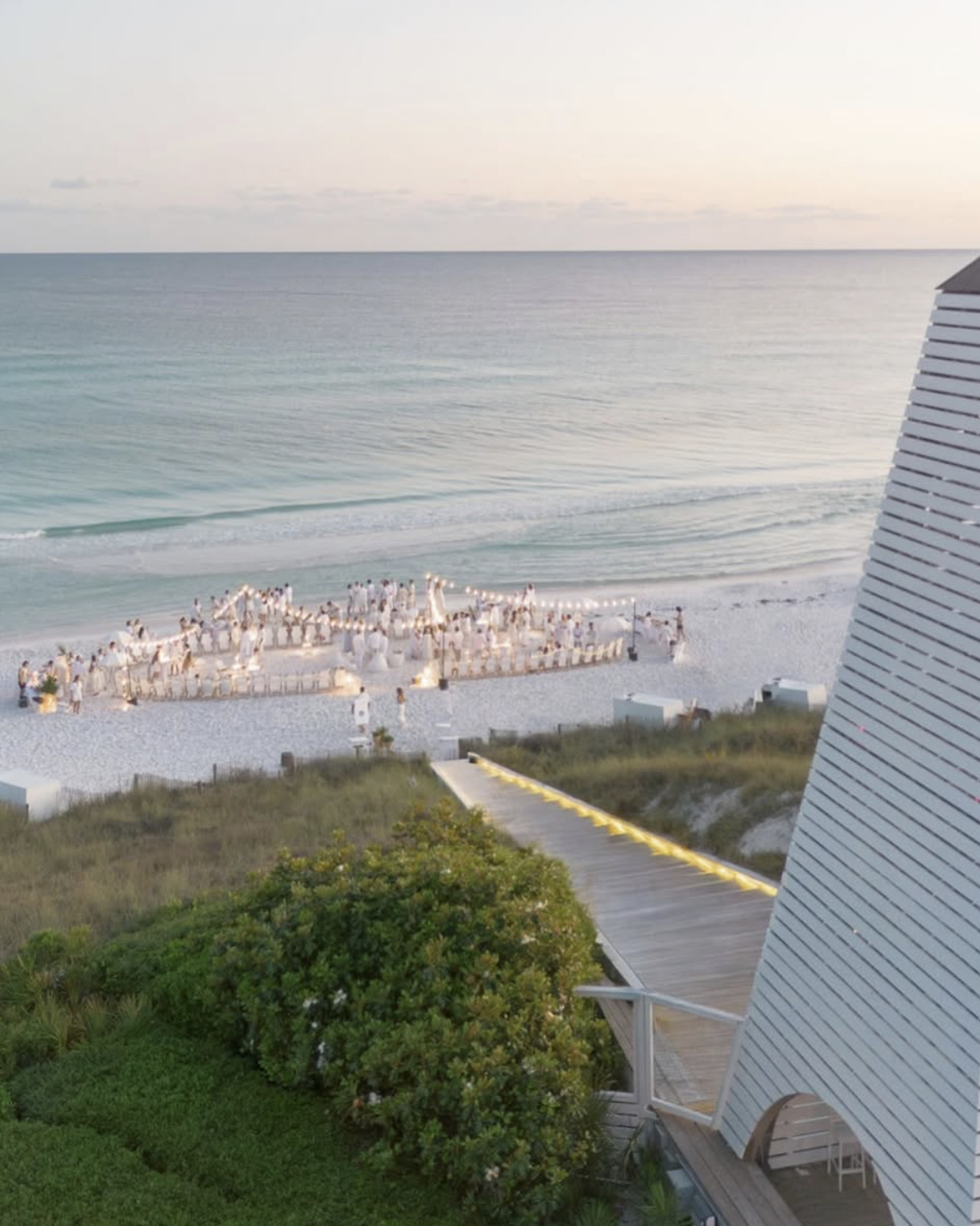 A beach scene with a large group of people dressed in white, forming a circle on the sandy shore, possibly for a wedding or event. The ocean waves are gently coming in, and the sky is light with soft colors, suggesting either sunrise or sunset. Part of a building with striped siding and some greenery are visible in the foreground.