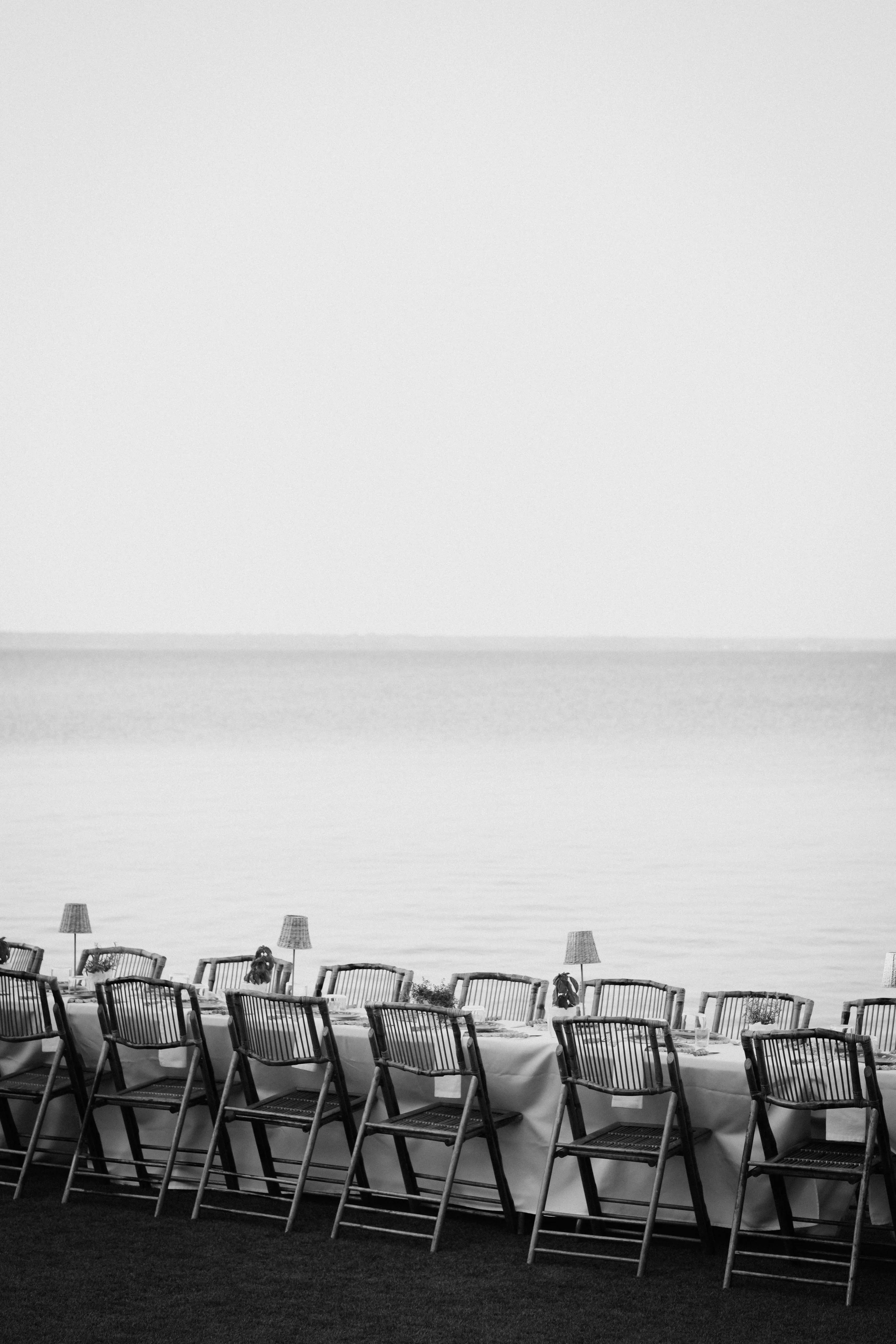 A black and white photo of a seaside outdoor dining setup with multiple chairs arranged along a long table covered with a cloth. There are lamps and small flower arrangements on the table, facing towards the water in the background, with calm sea and a distant horizon visible.