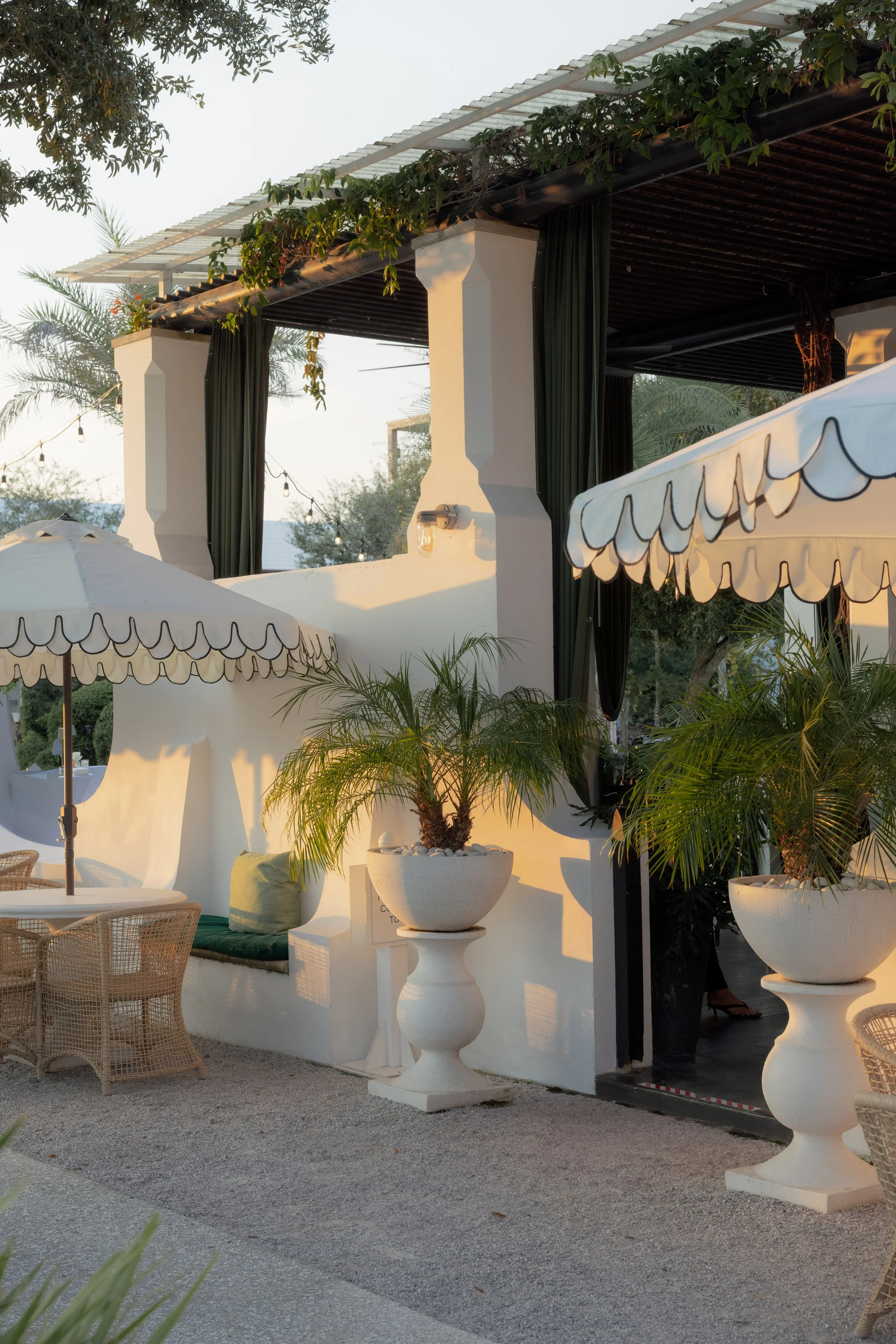 An outdoor patio area with white decorative planters and furniture, umbrellas, and lush greenery. The setting appears to be a relaxed, elegant space, possibly at a restaurant or resort, with warm lighting and a cozy atmosphere.