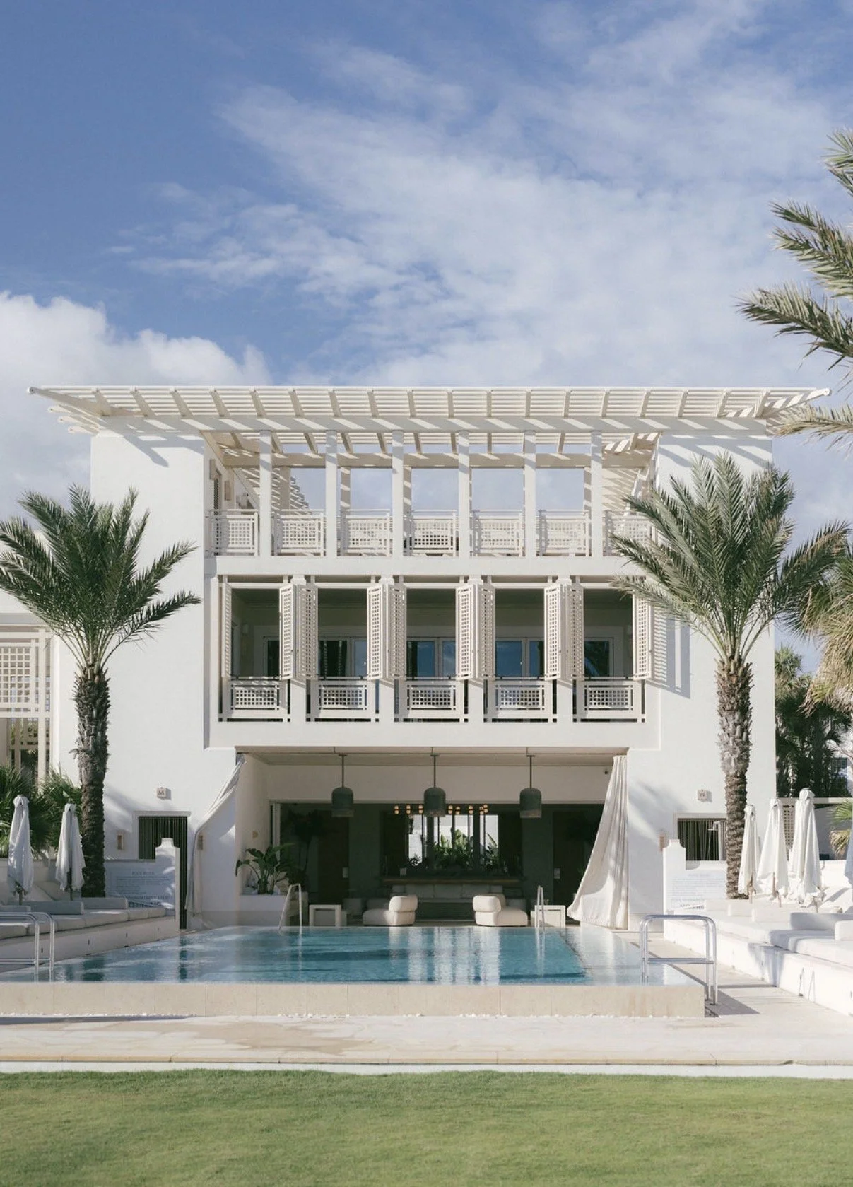 White modern building with multiple balconies and large windows, palm trees on either side, swimming pool in the foreground, with a blue sky and clouds above.