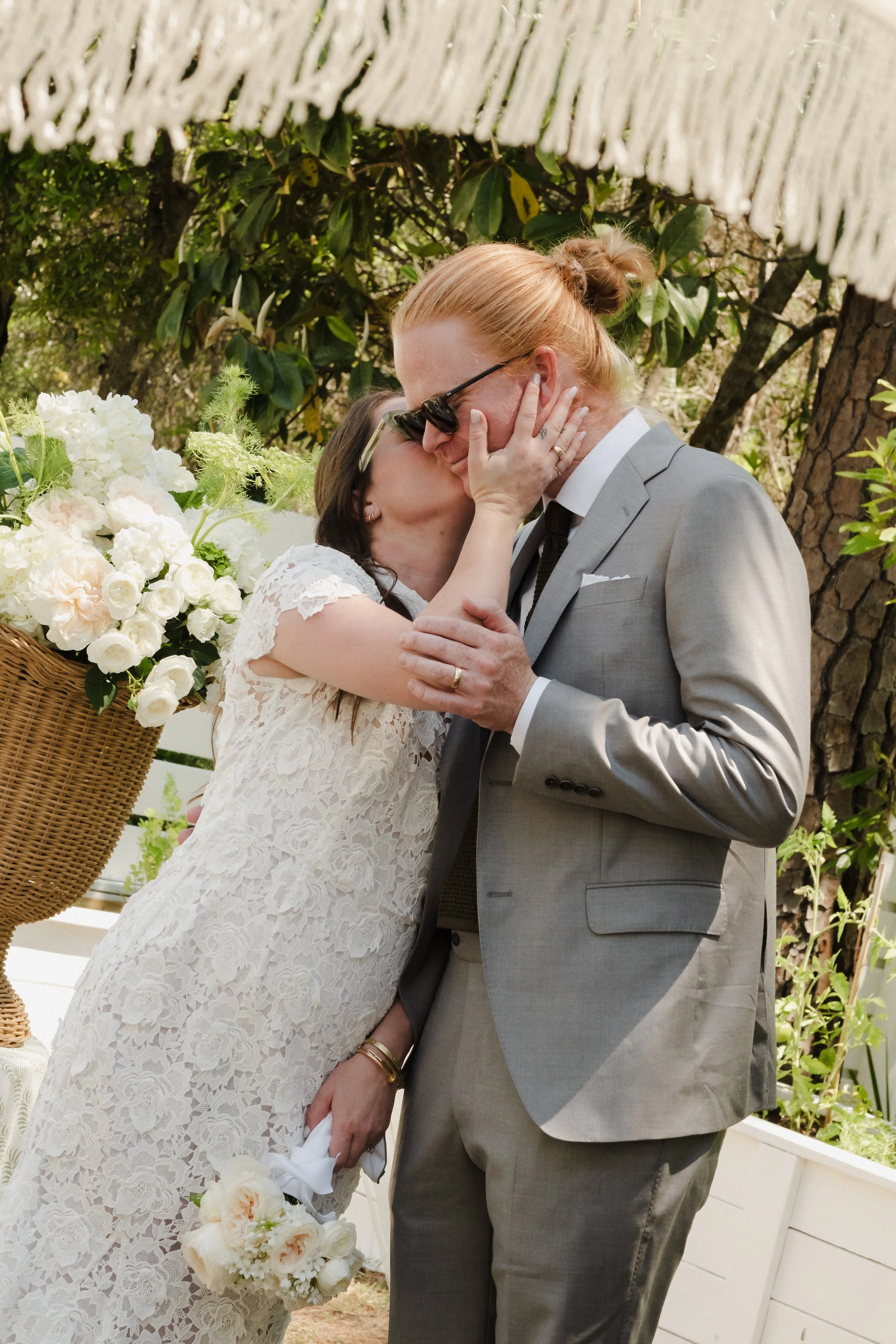 A bride and groom share a kiss during their wedding ceremony outdoors surrounded by trees and greenery. The bride wears a lace wedding dress and holds a bouquet of white flowers, while the groom wears a light gray suit and sunglasses.