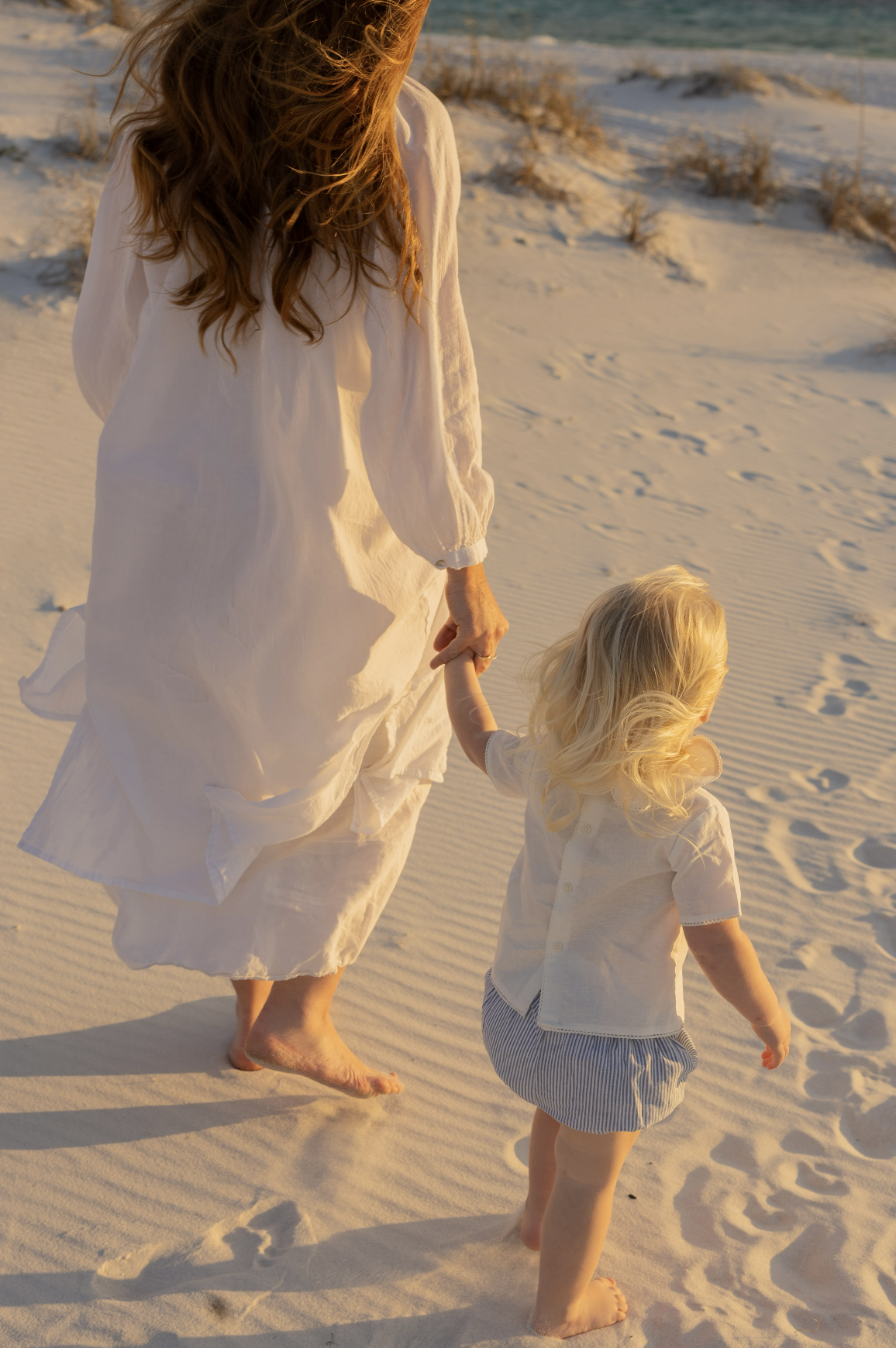A woman and a young boy walk barefoot on sandy beach at sunset, holding hands, with ocean and sand dunes in the background.