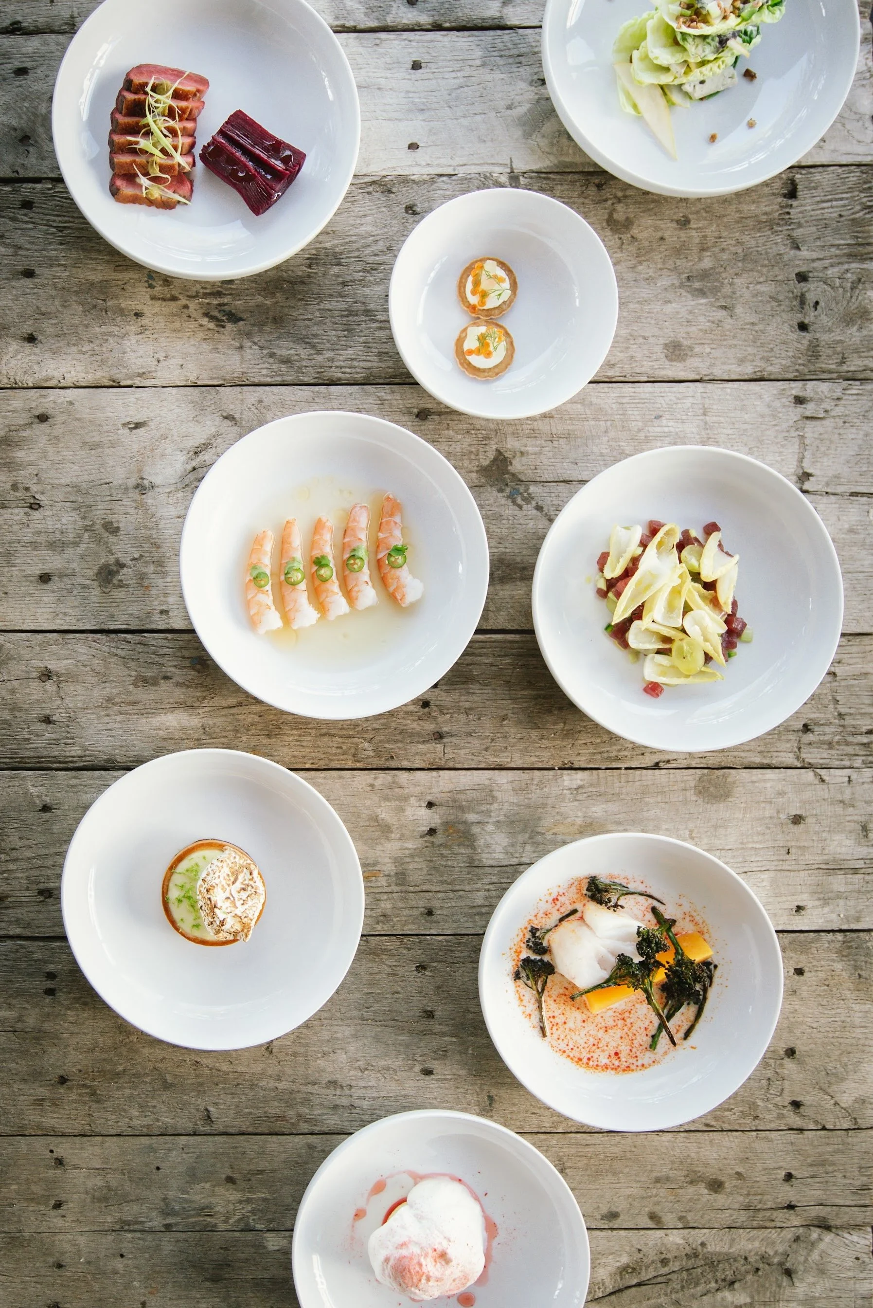An overhead view of several white plates with various gourmet dishes on a rustic wooden table.