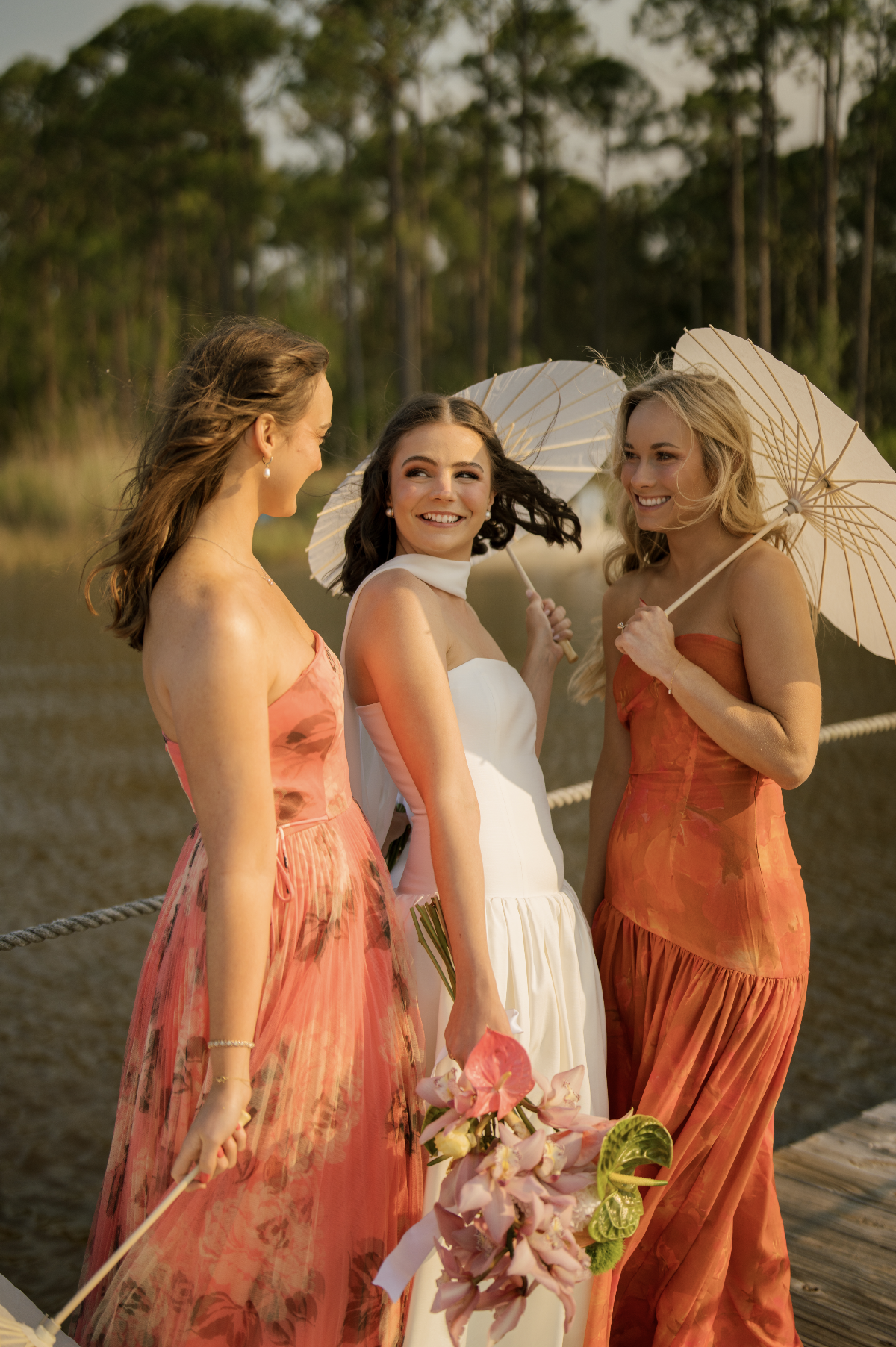 Three women dressed in colorful summer dresses standing on a dock near water, holding parasols and flowers, engaged in conversation and smiling, with a background of trees and a cloudy sky.