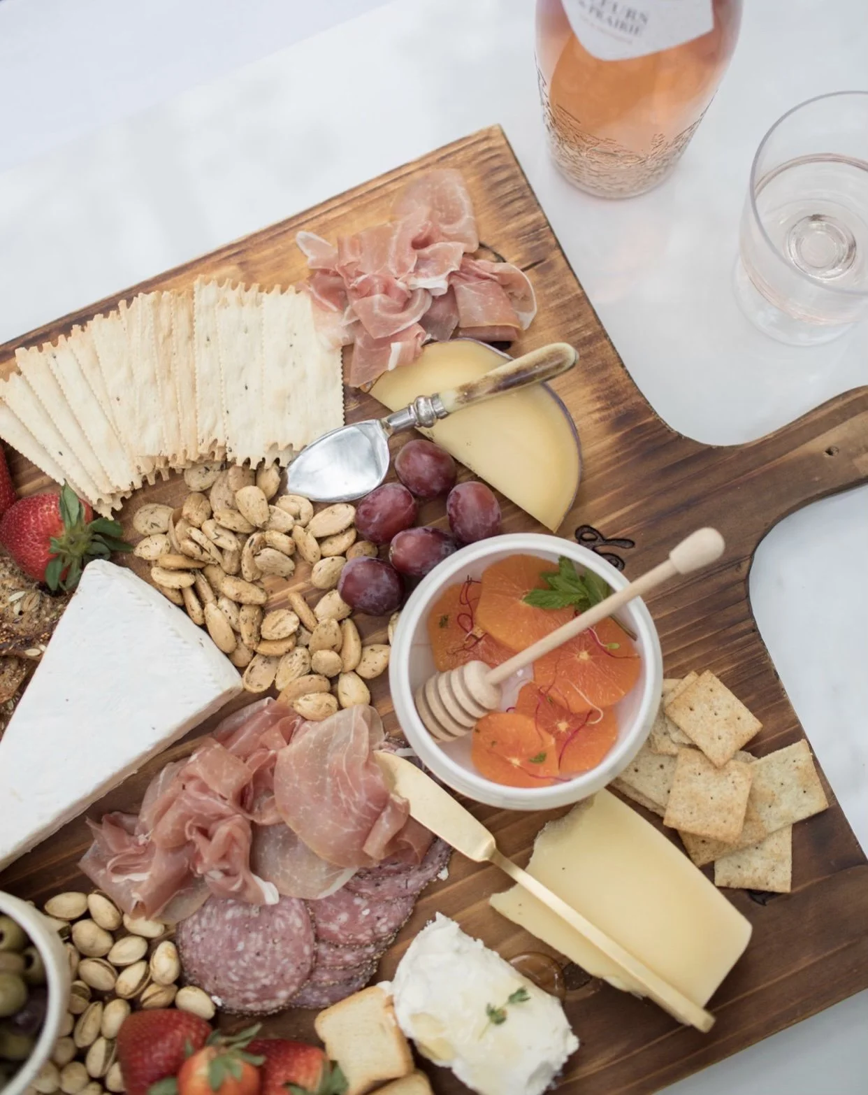 Charcuterie board with cheese, prosciutto, almonds, crackers, grapes, strawberries, honey, and toppings.