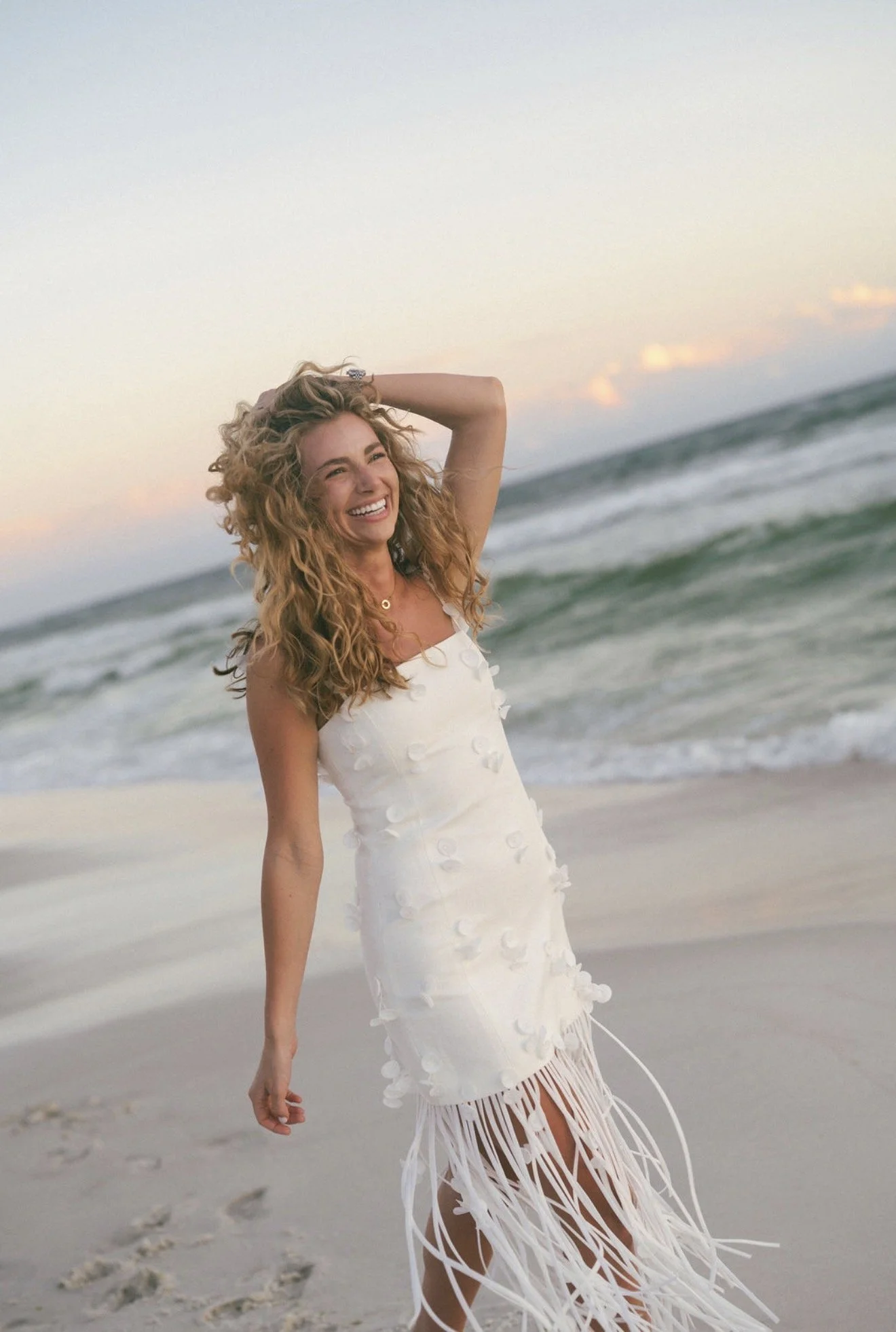 A woman with curly blonde hair smiling at the beach, wearing a sleeveless white dress with fringe at the bottom, with the ocean and sky in the background during sunset.