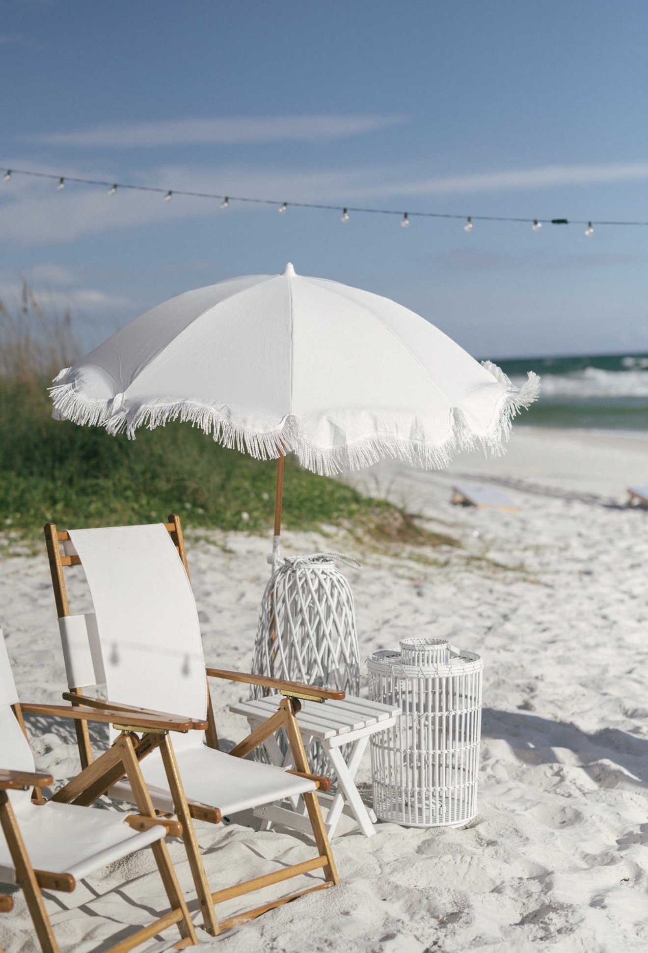 Beach scene with a white umbrella, wooden lounge chairs, and decorative white lanterns on white sand, with ocean waves and a blue sky in the background.