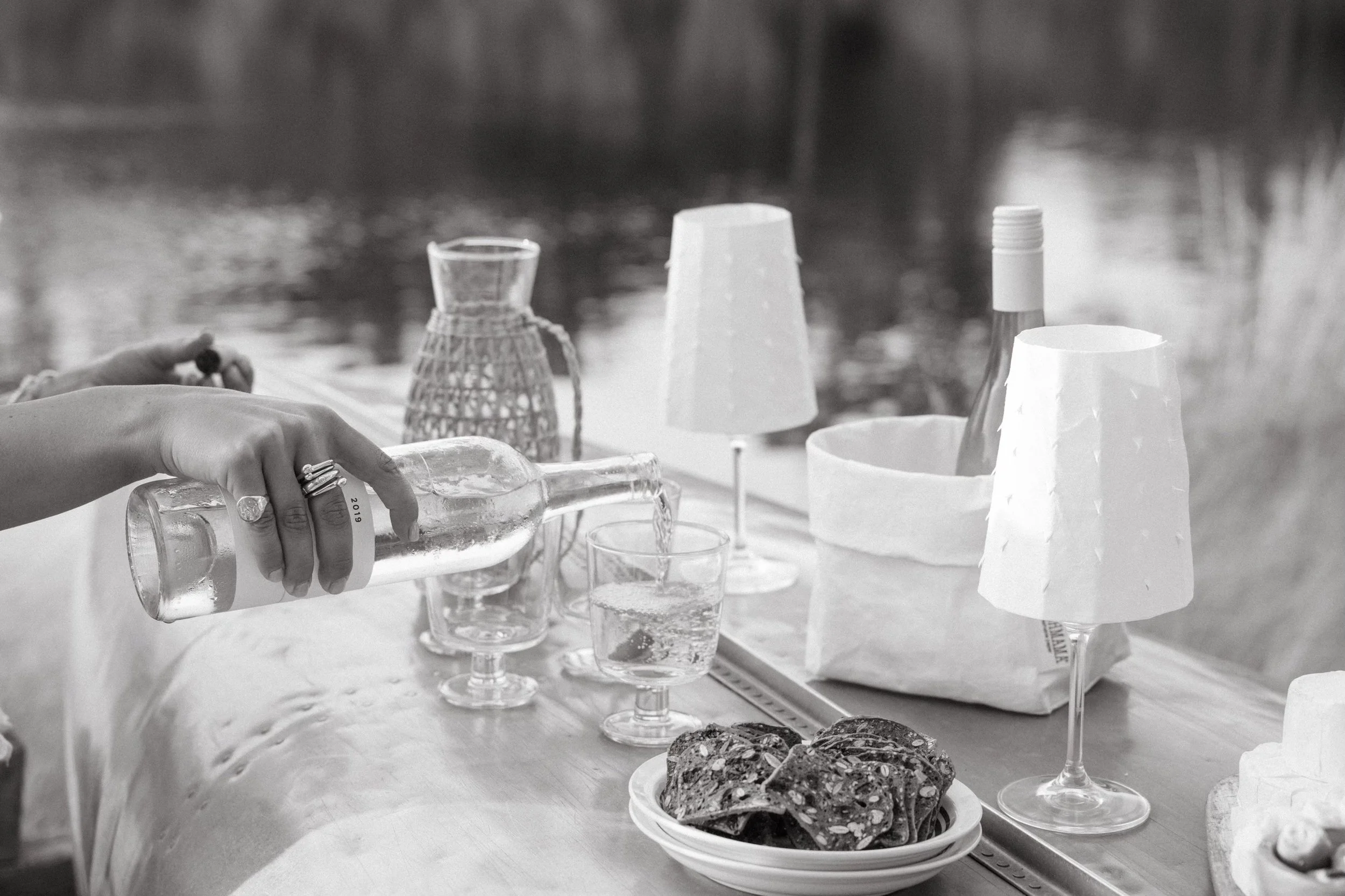 A person is pouring water from a glass bottle into a glass cup on a table. The table also has several other glasses, paper lanterns, and a plate of cookies. The background appears to be a blurred outdoor scene.