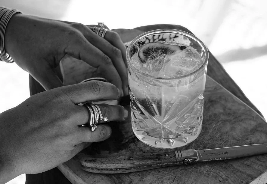 A person's hands with rings on fingers holding a glass of water with ice and lemon, on a wooden tray with a metal straw.