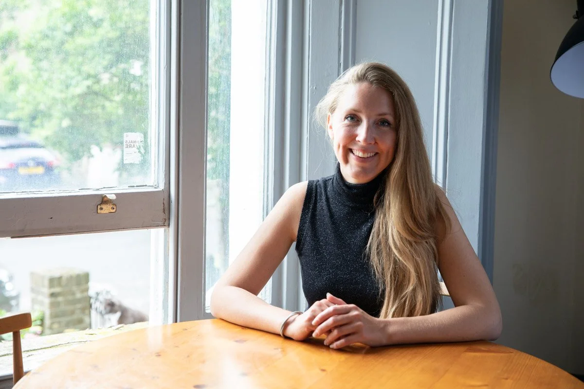 A young woman with long blonde hair smiling and sitting at a wooden table by a large window with green trees outside.