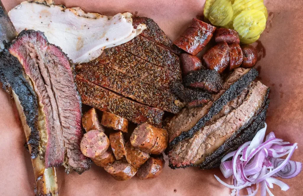 Assorted smoked barbecue meats including brisket, sausage, and ribs, served with pickles and sliced onions.