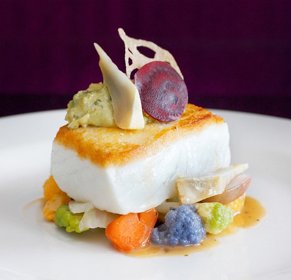 Pan-seared white fish fillet topped with white chocolate, a dollop of mashed potatoes with herbs, a small beet slice, a white chocolate lattice, and colorful vegetable puree with sauce on a white plate.
