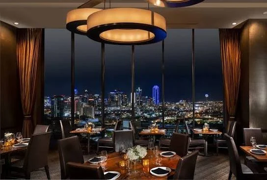 Interior view of a restaurant with a city skyline at night visible through large windows. The dining tables are set with glasses, plates, and cutlery, and there are decorative centerpieces. Overhead lighting provides ambient illumination.