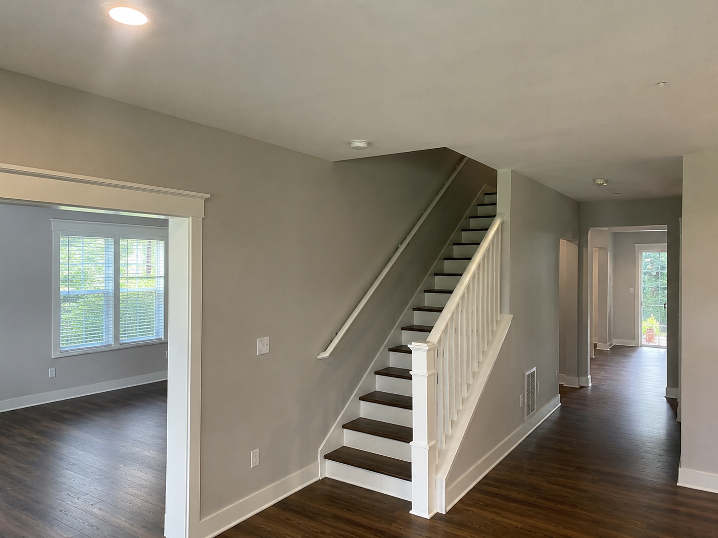 Interior view of a modern home with a staircase featuring dark wood steps, white risers, a white handrail, and spindles. The space has light gray walls, hardwood flooring, and large windows with white trim, allowing natural light in.