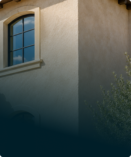 Close-up of a beige stucco house wall with a large arched window and a small bush in the foreground.