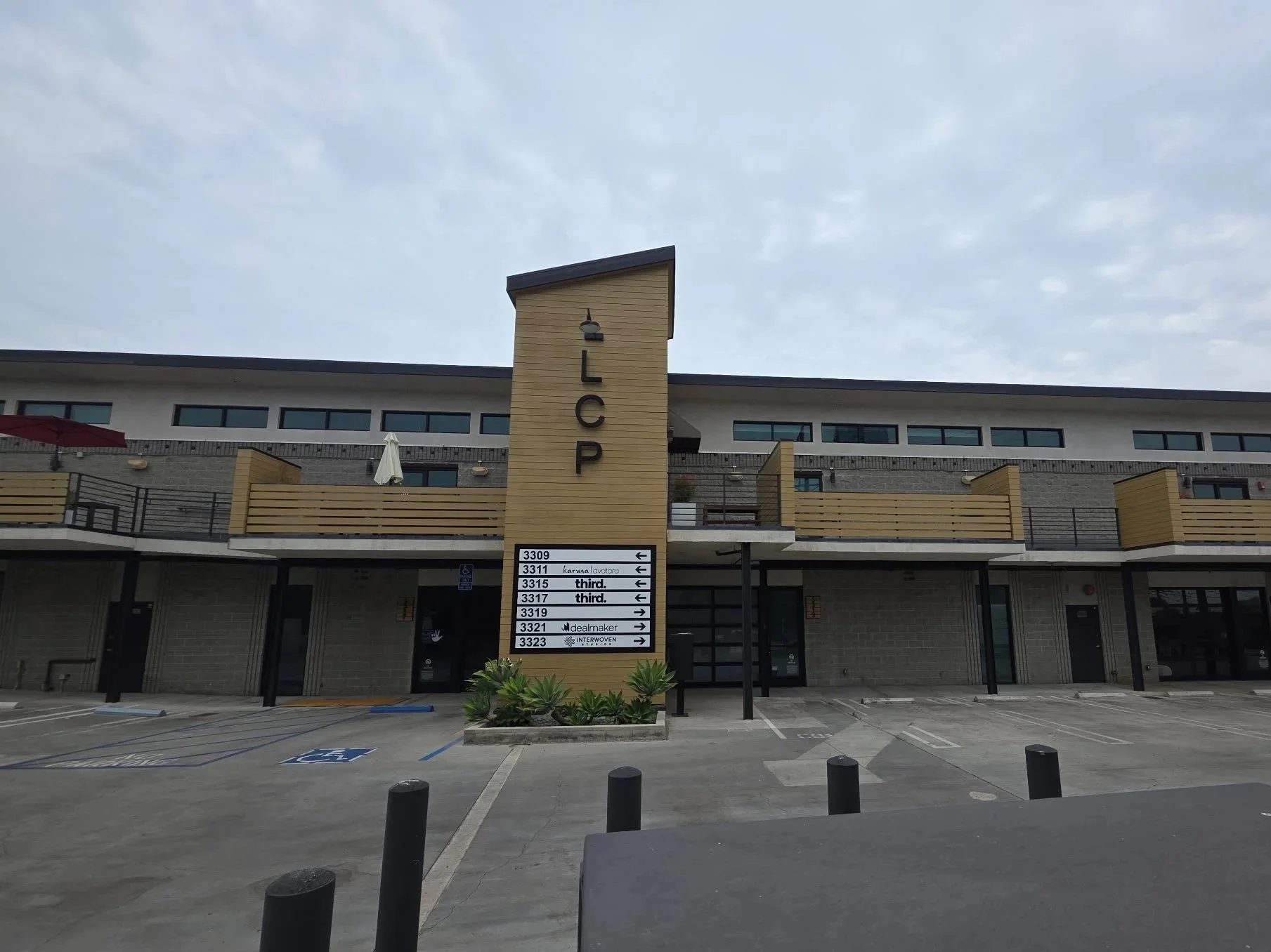 A modern multi-story building with a beige and gray exterior, featuring a sign with various business names and directions. There are parking spaces in front, including a designated handicapped spot, and a small landscaped area with plants. The sky is