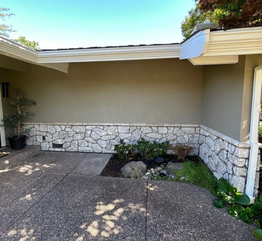 A house exterior with a stone facade, a small garden bed, pebbles, and a potted plant, under a clear blue sky.