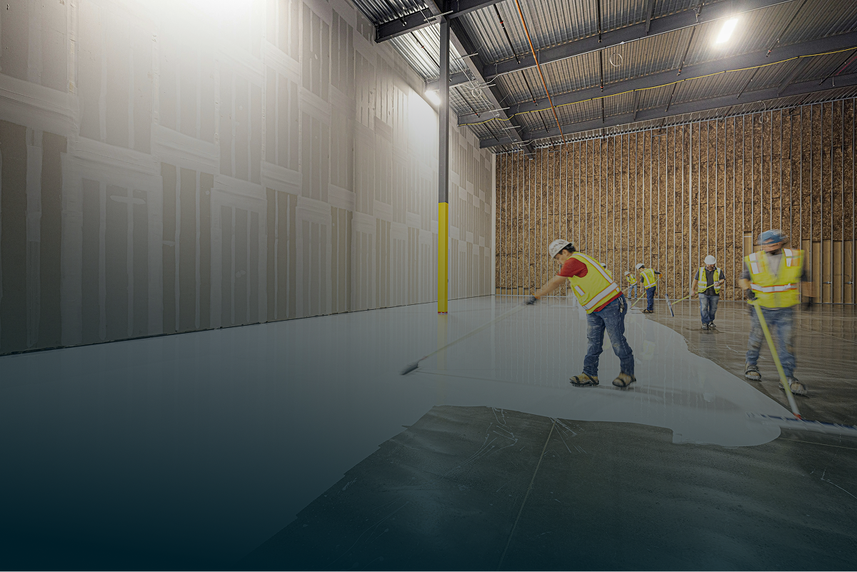Construction workers wearing safety vests and helmets installing a large white epoxy floor in an industrial warehouse.