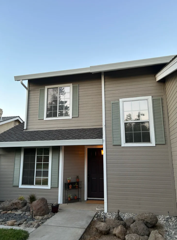 Two-story beige house with white and green shutters on the windows, a black front door, and a small front yard with rocks and plants.