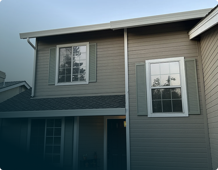 Front of a two-story house with beige siding and white trim, featuring two large windows with shutters and a dark front door.