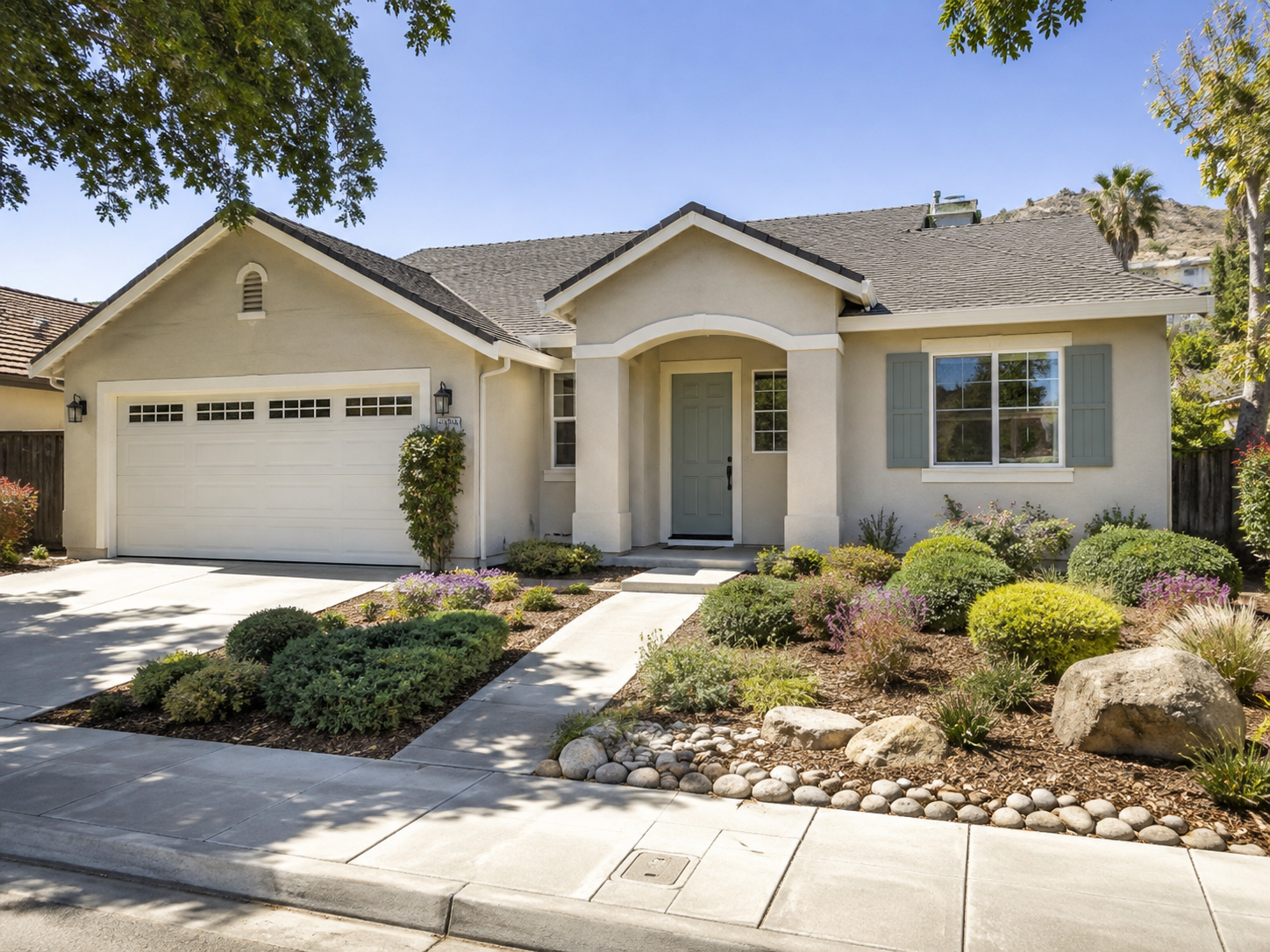 Front view of a single-story house with a white garage door, a small front porch, and landscaped yard with bushes and rocks, under a clear blue sky.
