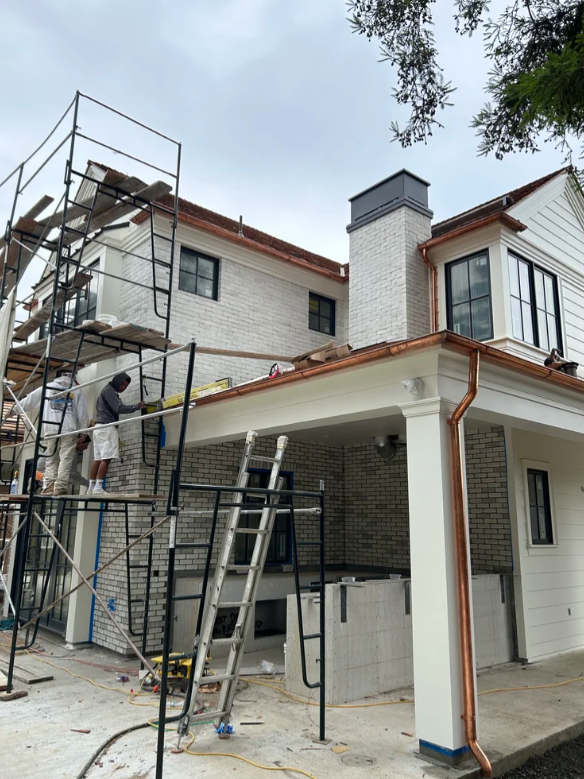 Construction workers on scaffolding working on the exterior of a house with brick and white siding, with ladders and tools visible.