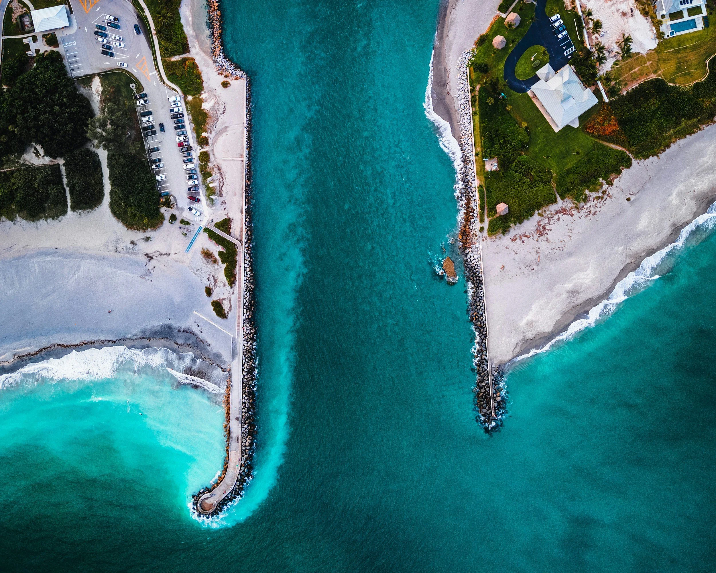 Aerial view of a jetty extending into the ocean with turquoise waters, surrounded by rocky breakwaters and lush green landscape, including a white building and parked cars.