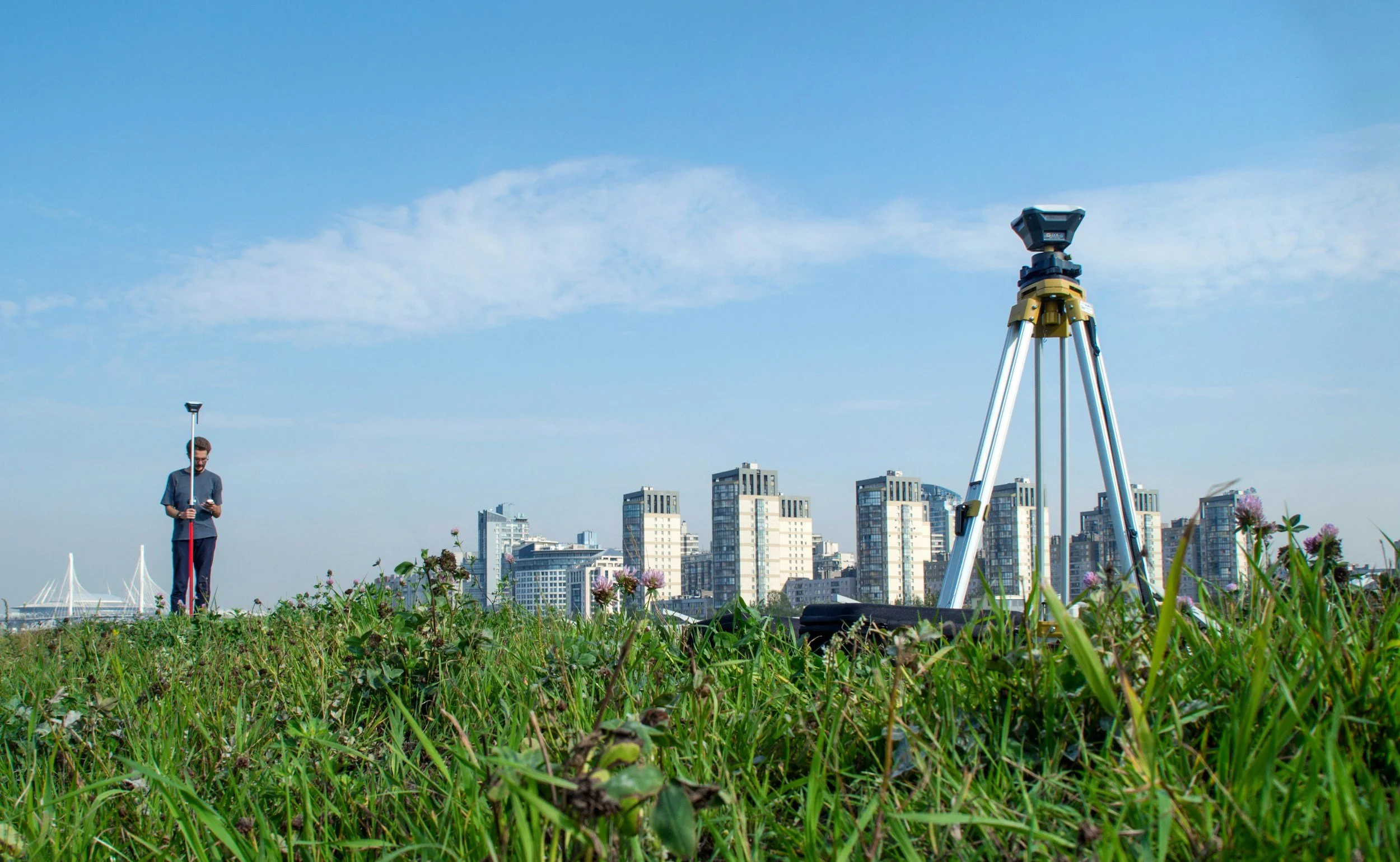 A person standing in a grassy field holding a surveying instrument, with a tripod set up nearby, in front of a cityscape with tall buildings under a blue sky.