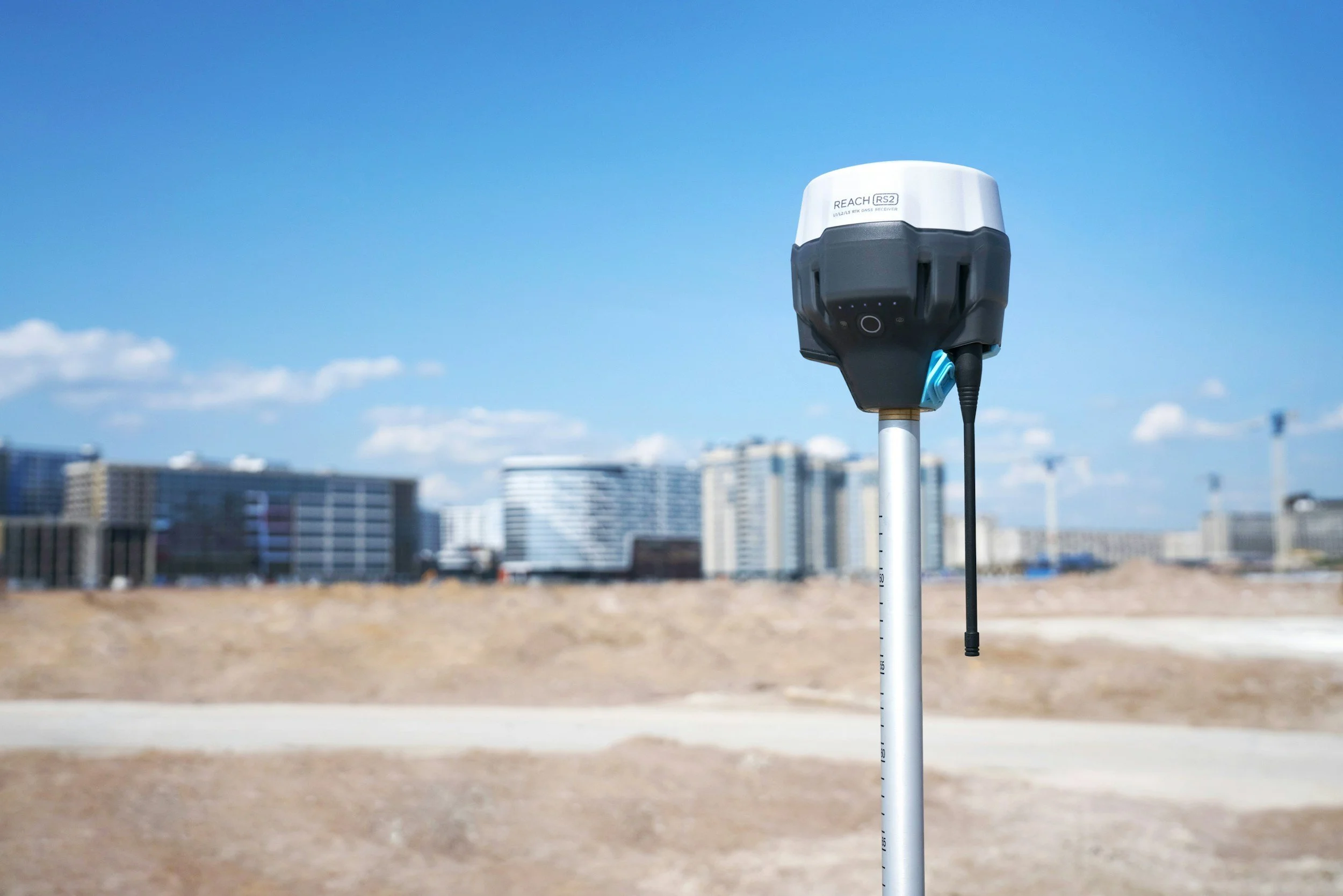 A weather station or sensor mounted on a pole outdoors with a clear blue sky and city buildings in the background.