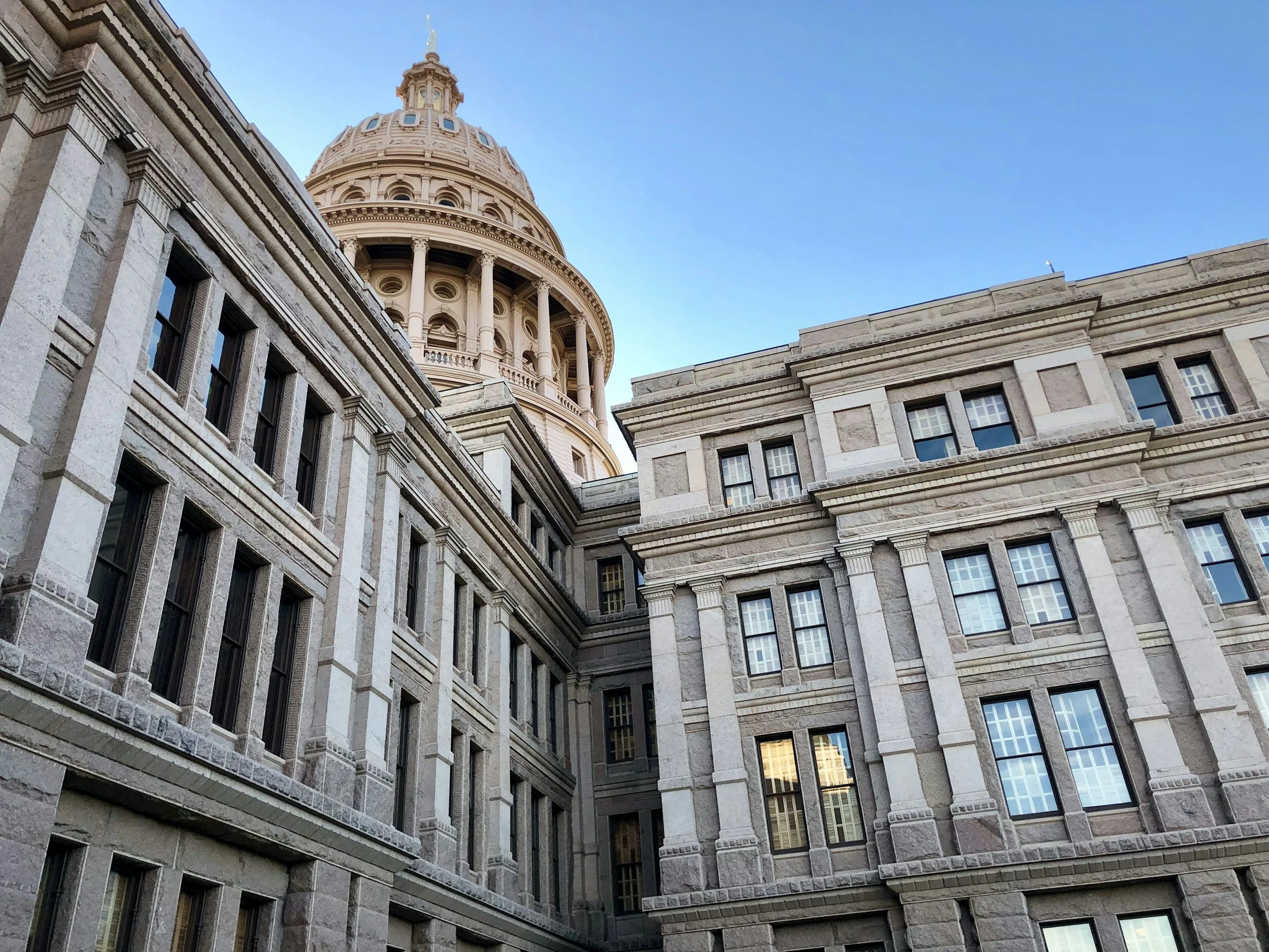Low-angle view of historic U.S. Capitol building with a prominent dome, surrounded by adjacent stone buildings and a clear blue sky.