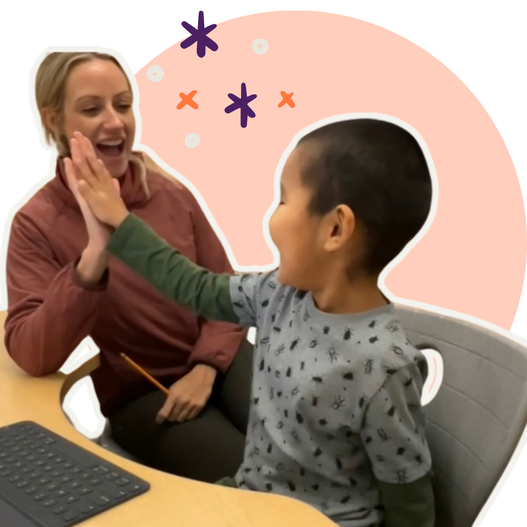 A young boy with short dark hair, wearing a gray shirt with black designs, giving a high five to a smiling woman with blonde hair, dressed in a pink jacket, sitting at a table with a computer keyboard nearby. The background has pink and purple decorative elements.