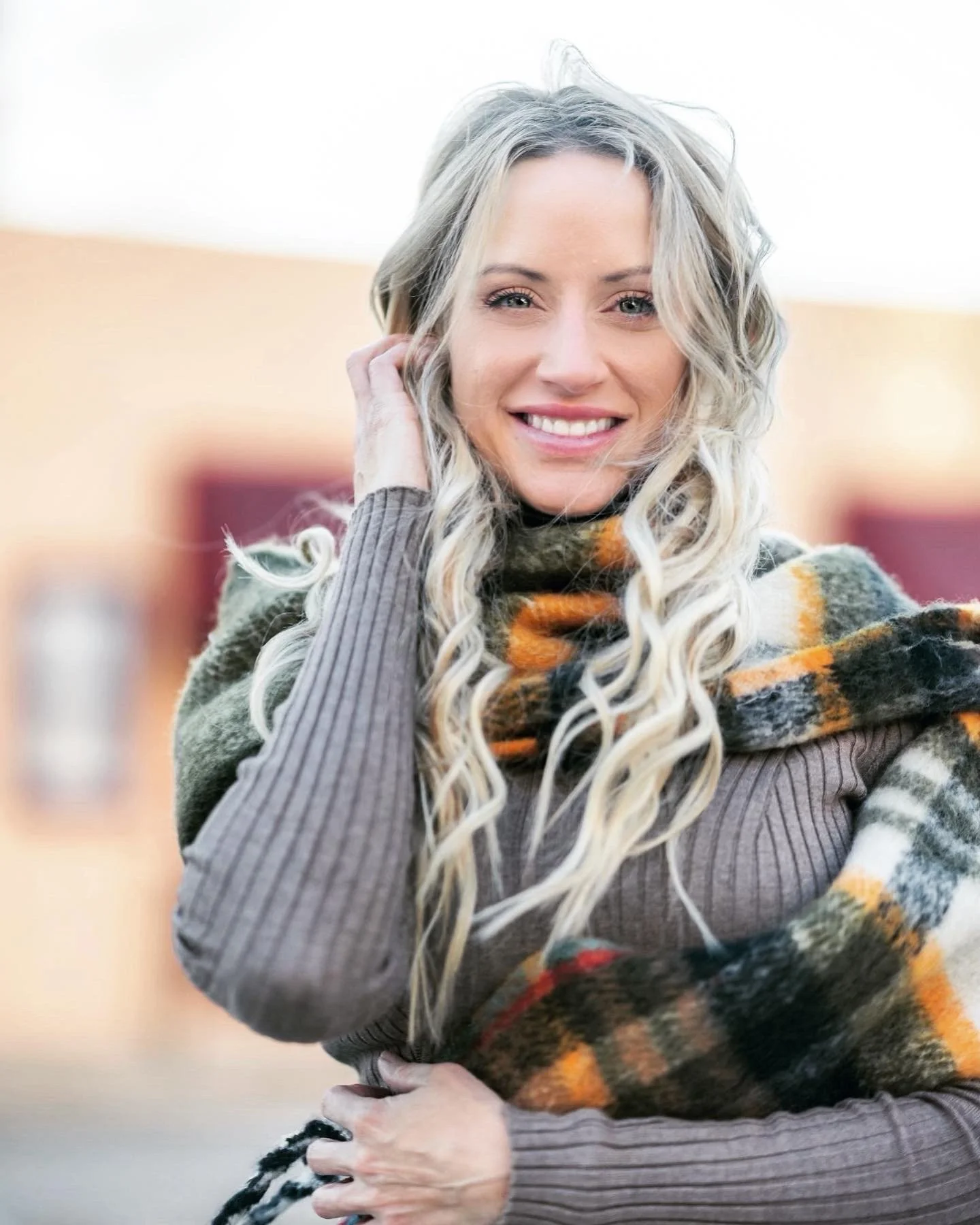 A woman with long, wavy gray hair, smiling, wearing a striped sweater and a multicolored plaid scarf.