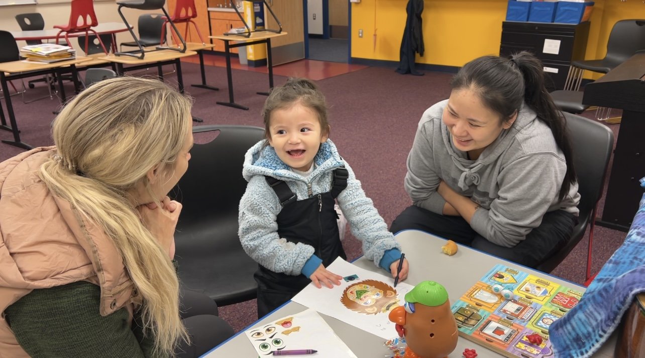 A young girl with a big smile, wearing a blue and black jacket, is sitting at a table with two women, one on each side. The woman on the left has long blonde hair and is wearing a beige jacket, while the woman on the right has dark hair tied back and is wearing a gray hoodie. The table has colorful toys, pictures, stickers, and drawing materials on it. The room has a maroon carpet, yellow and orange walls, and several chairs and tables in the background.