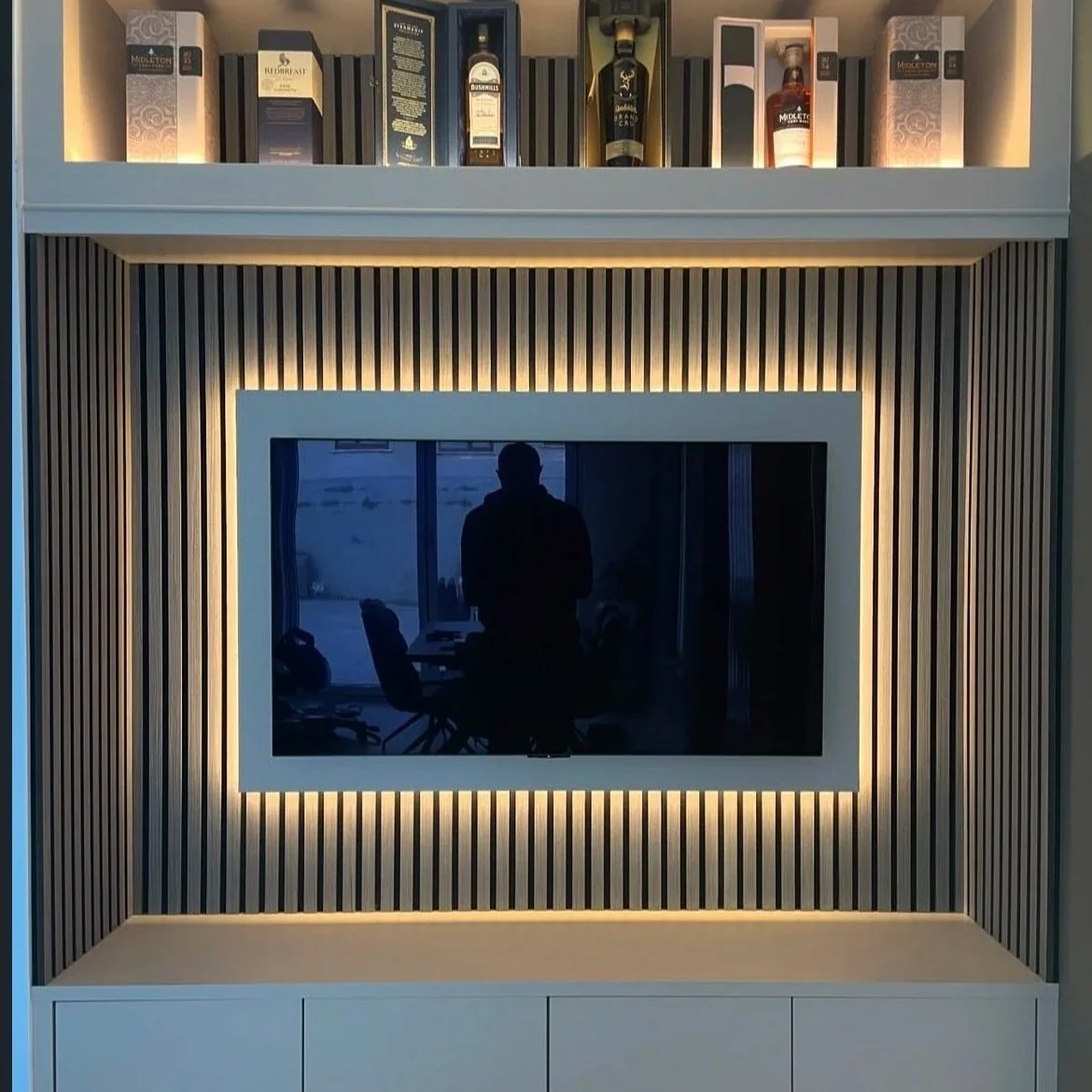 A modern entertainment center with a wall-mounted TV surrounded by vertical wooden slats and LED lighting. The top shelf displays various bottles of liquor.