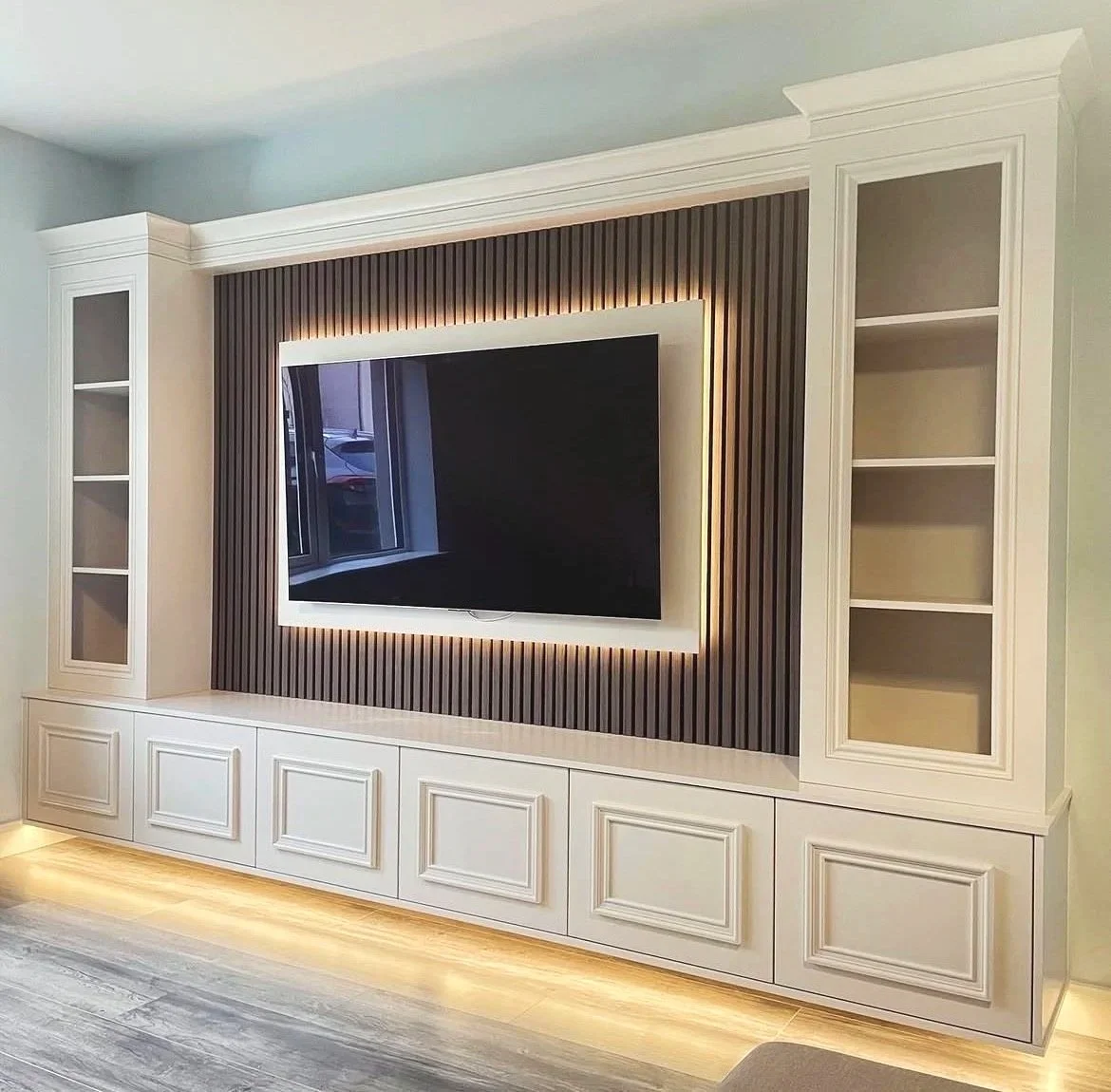 Modern white entertainment center with built-in shelving, a flat-screen TV on a textured wood-paneled wall with backlighting, and hardwood flooring.