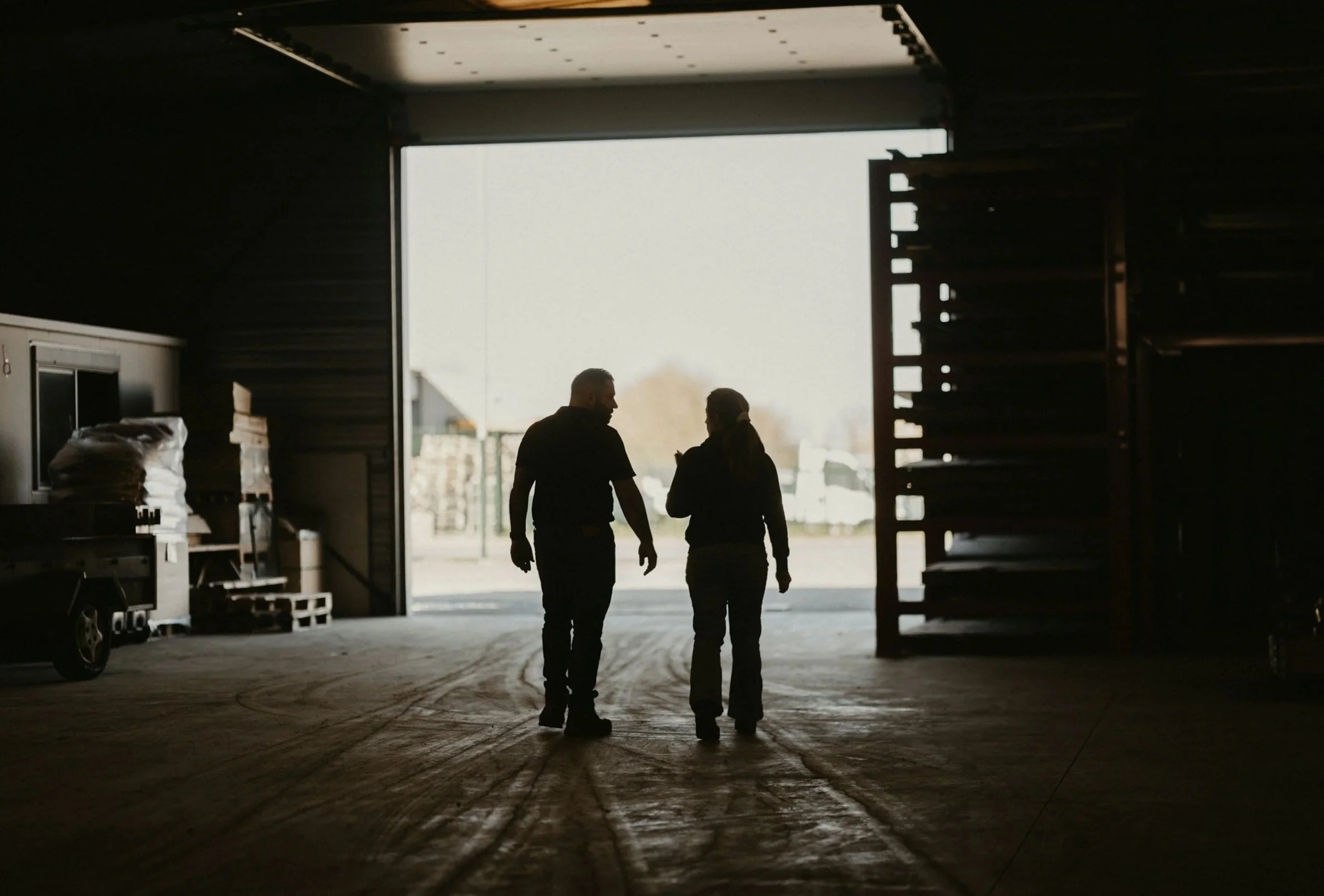 Two people walking inside a warehouse or industrial building, silhouetted against the bright open loading dock entrance.