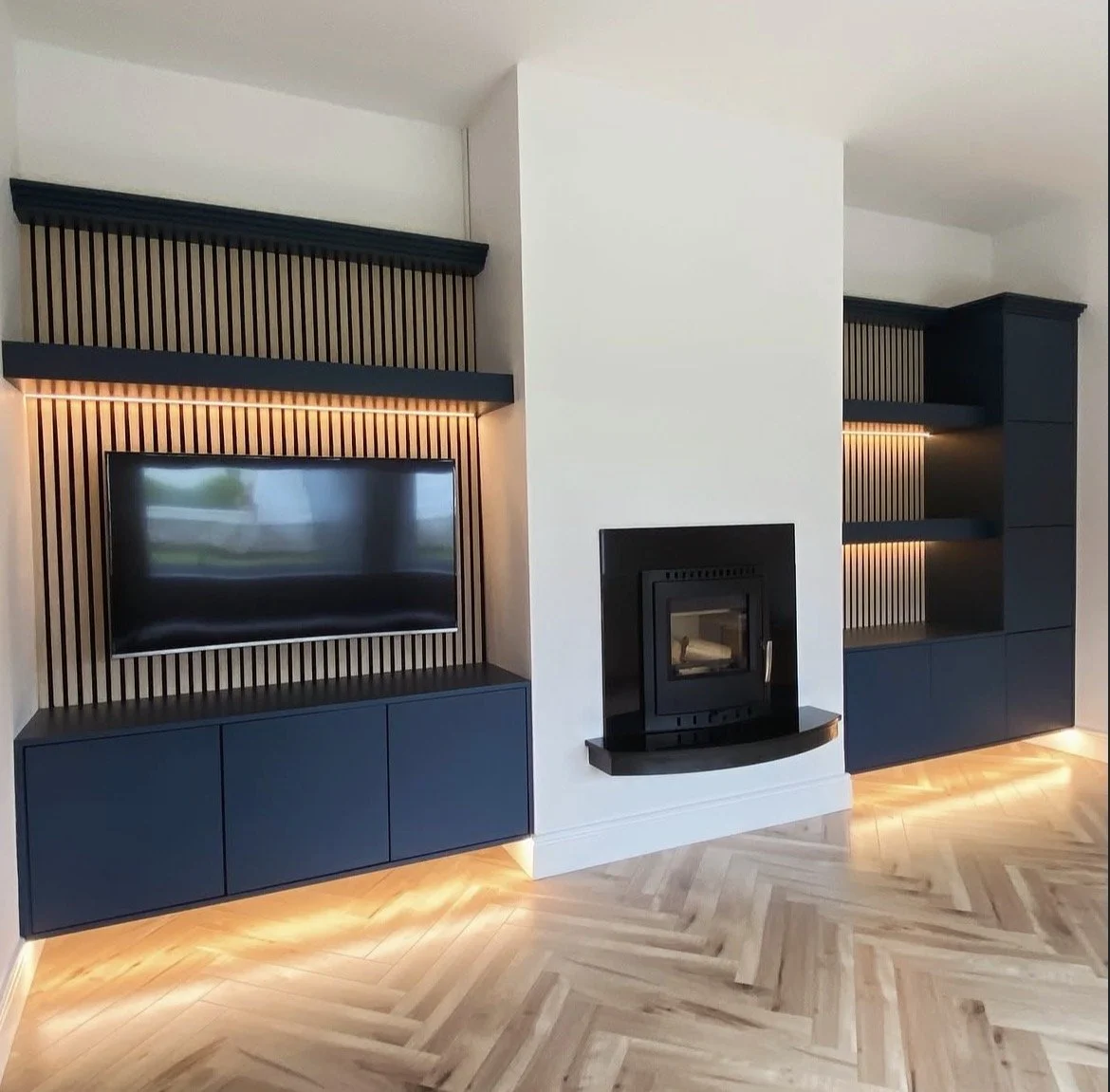 Modern living room features a flat-screen TV on a black cabinet with wood slats backdrop, a black fireplace, and built-in shelving with lighting, all on a herringbone wood floor.