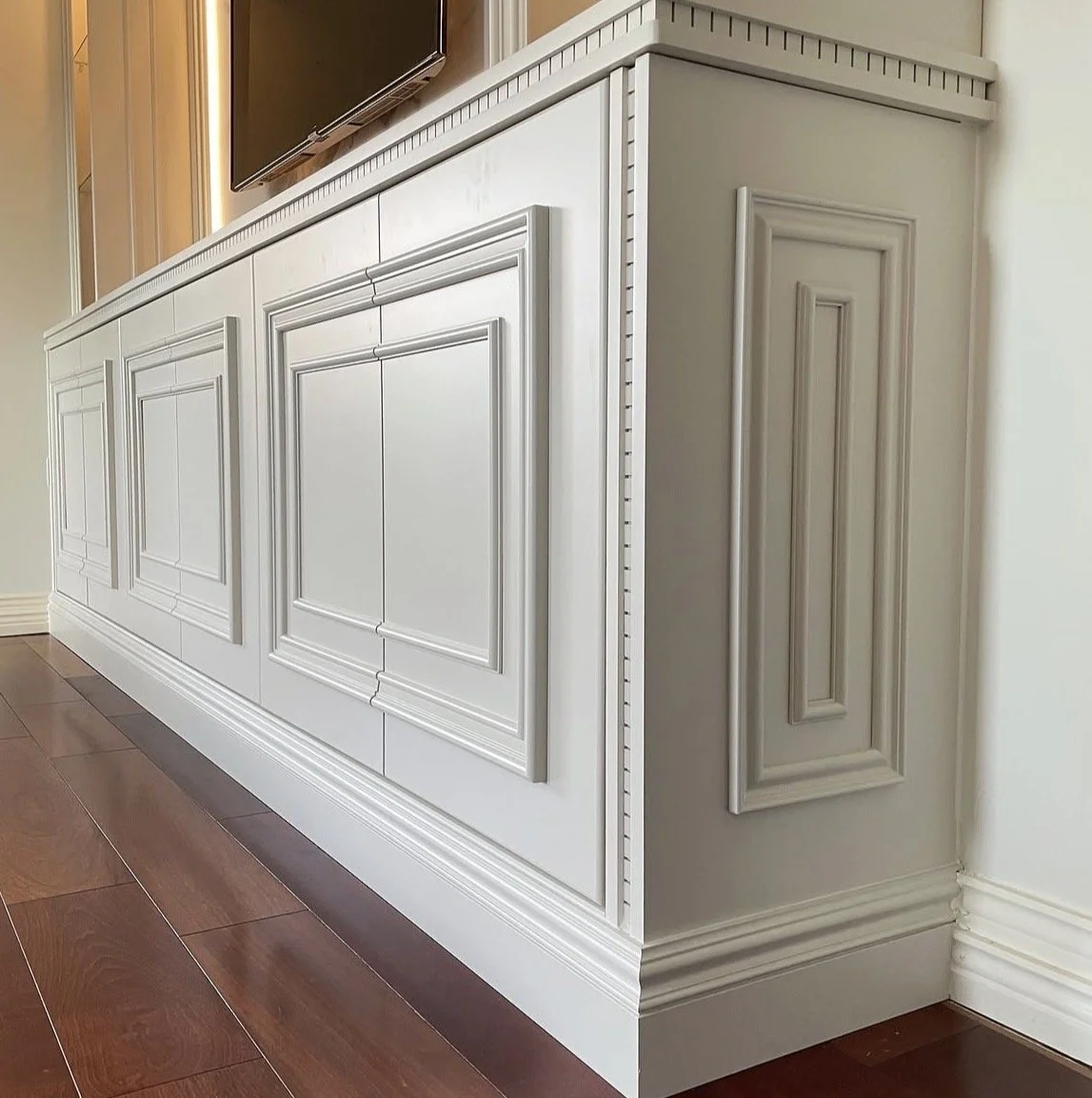 Close-up view of a white wooden cabinet with detailed molding and paneling, situated on a hardwood floor in a room.