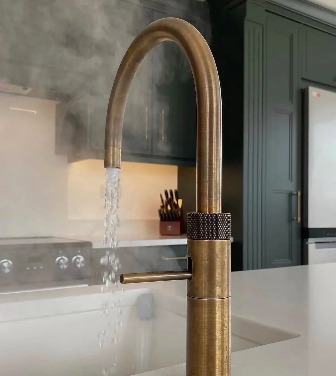 Brushed gold kitchen faucet with water running into a white sink, background shows dark green cabinets and a utensil holder.