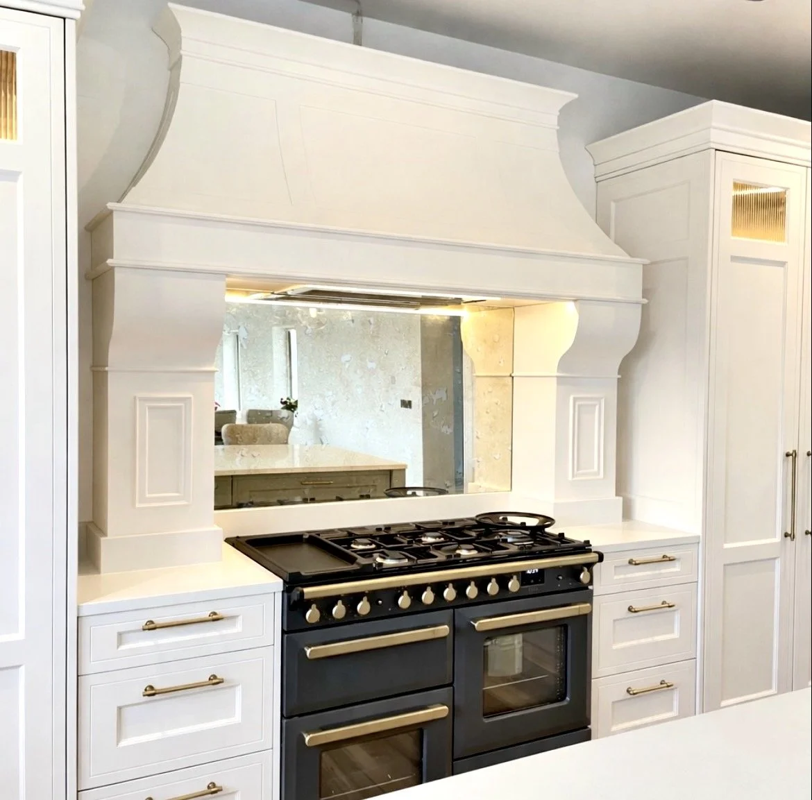 White kitchen range oven with a black cooktop, surrounded by white cabinetry with gold handles and decorative molding, reflective backsplash or mirror behind it.