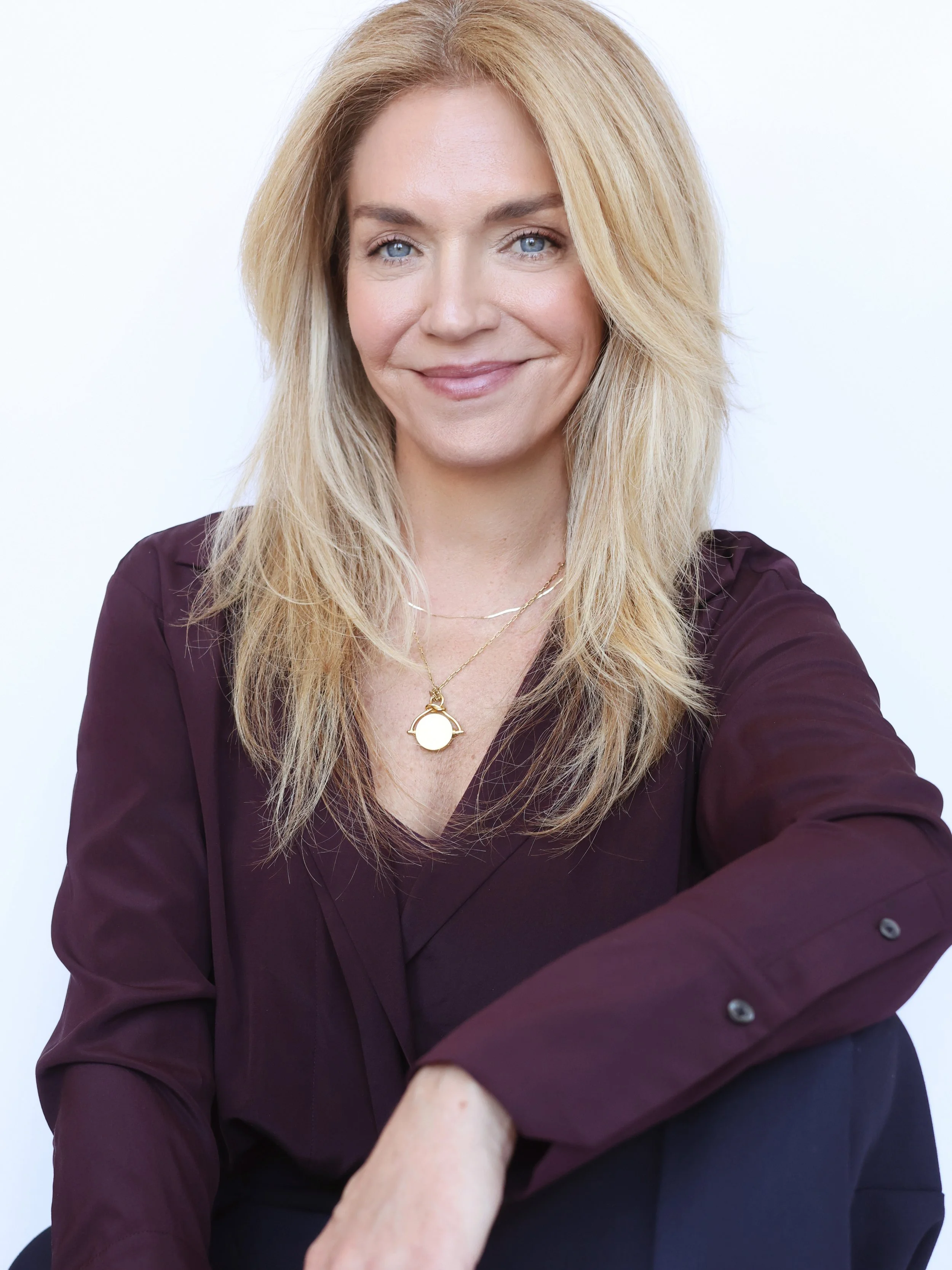 A woman with blonde hair and blue eyes smiling, wearing a dark maroon blouse and a gold necklace with a circular pendant, sitting with her arms crossed.