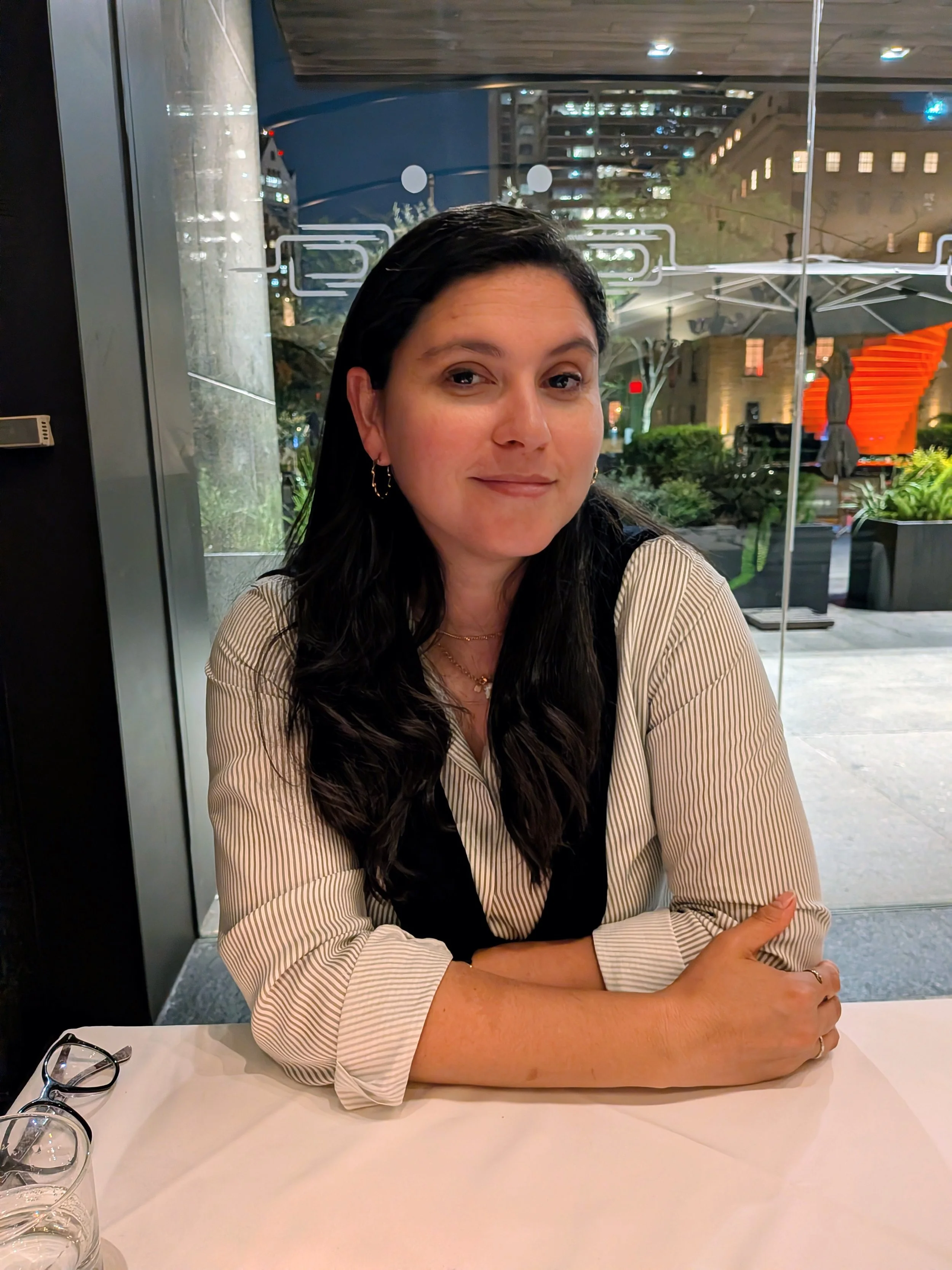 A woman sitting at a table in a restaurant or cafe, with arms crossed on the table, looking at the camera, wearing a striped shirt and black vest, with glasses on the table and city buildings visible through a large window behind her.