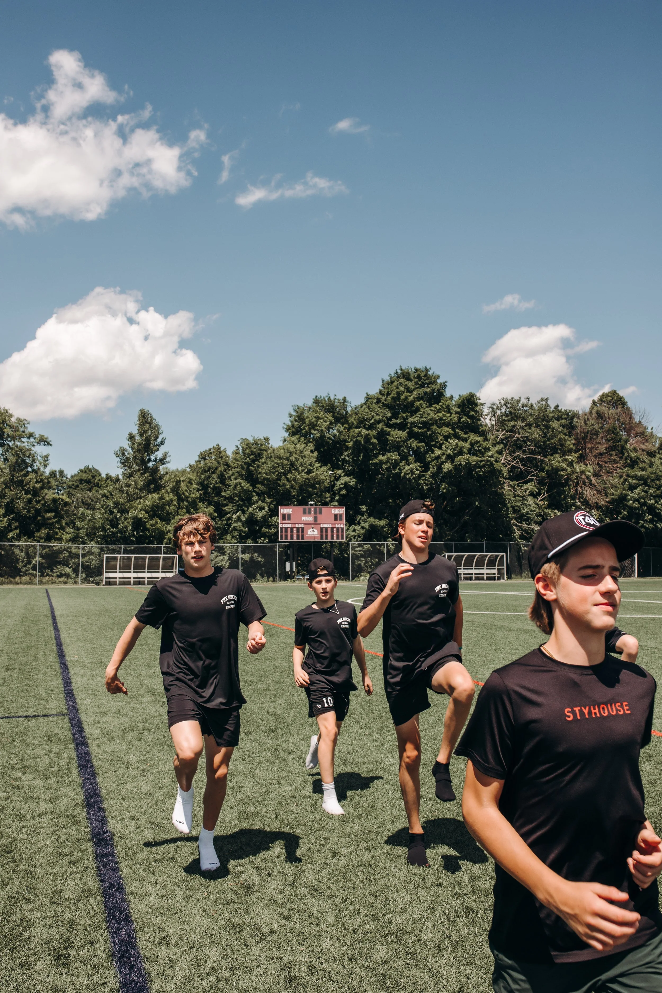 A group of young people running on a sports field under a clear blue sky with some clouds, with a scoreboard and green trees in the background.