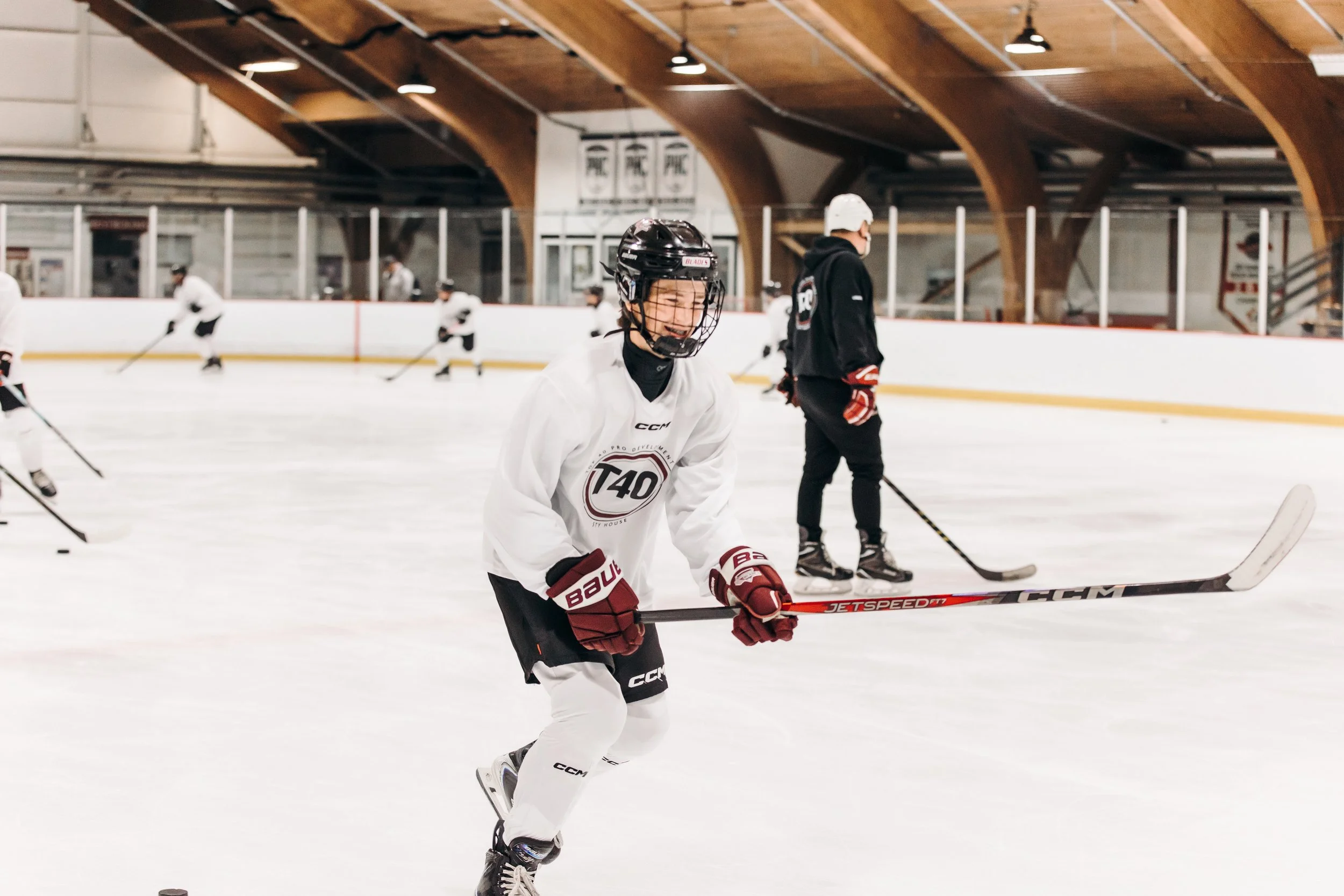 Young hockey player in white jersey with a black helmet smiling on ice rink during practice, with other players and a coach visible in the background.