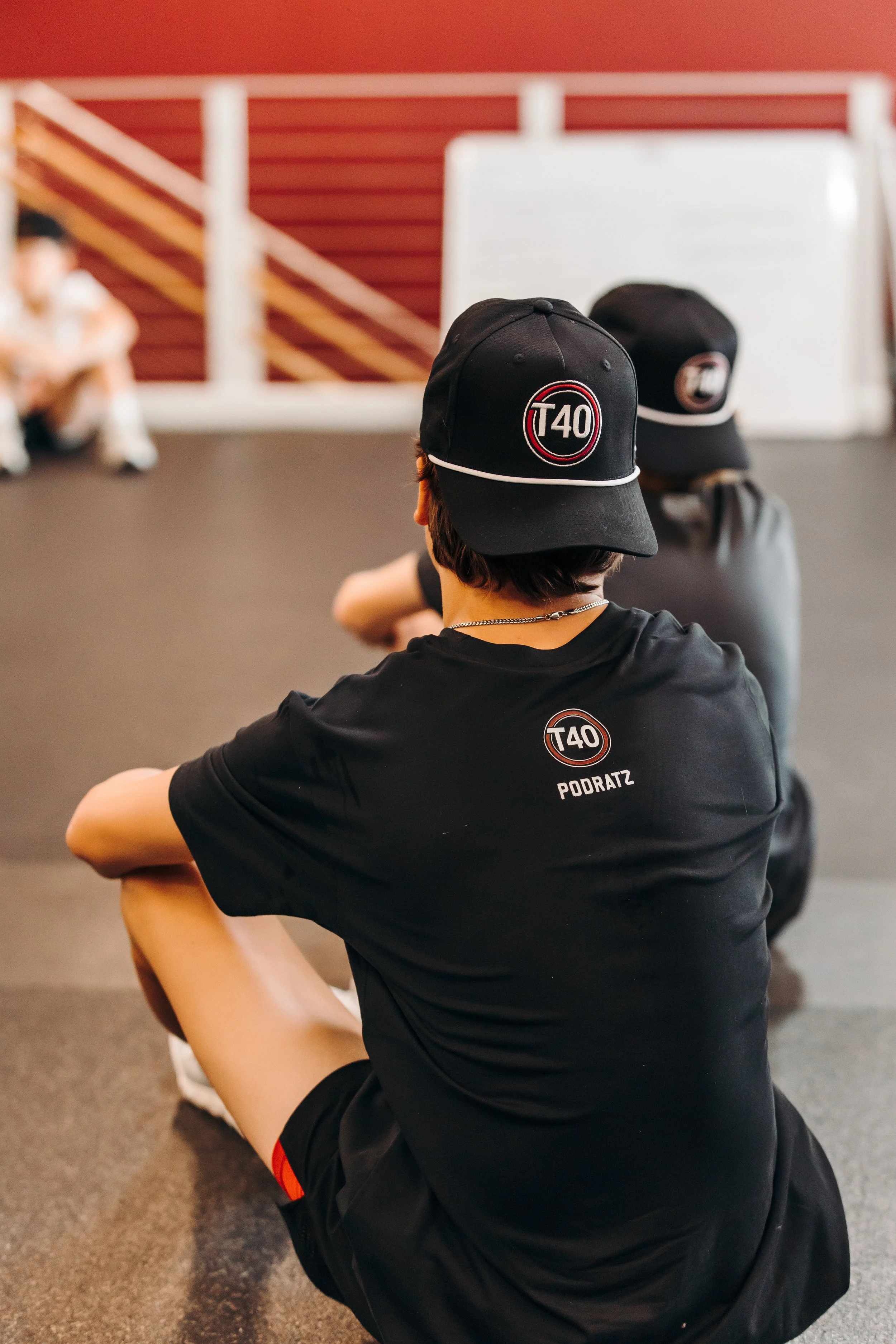 A person wearing a black T40 cap and T40 shirt sitting on the floor, with their arms resting on their knees, in a room with a staircase and a whiteboard in the background.