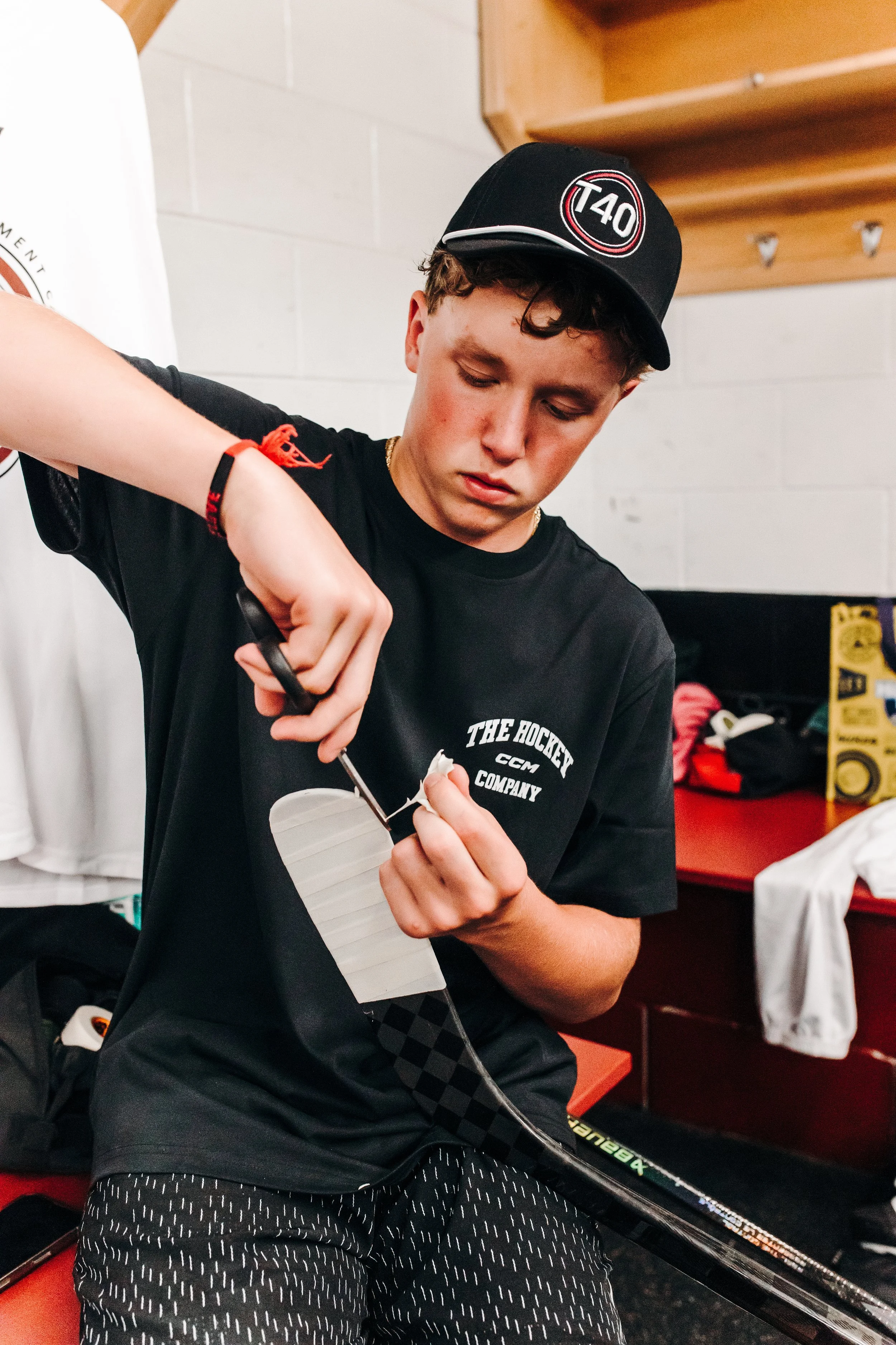 A young male hockey player with curly hair, wearing a black cap with the text "T40" and a black T-shirt with white text, is preparing his hockey stick in a locker room with white brick walls and wooden shelves.