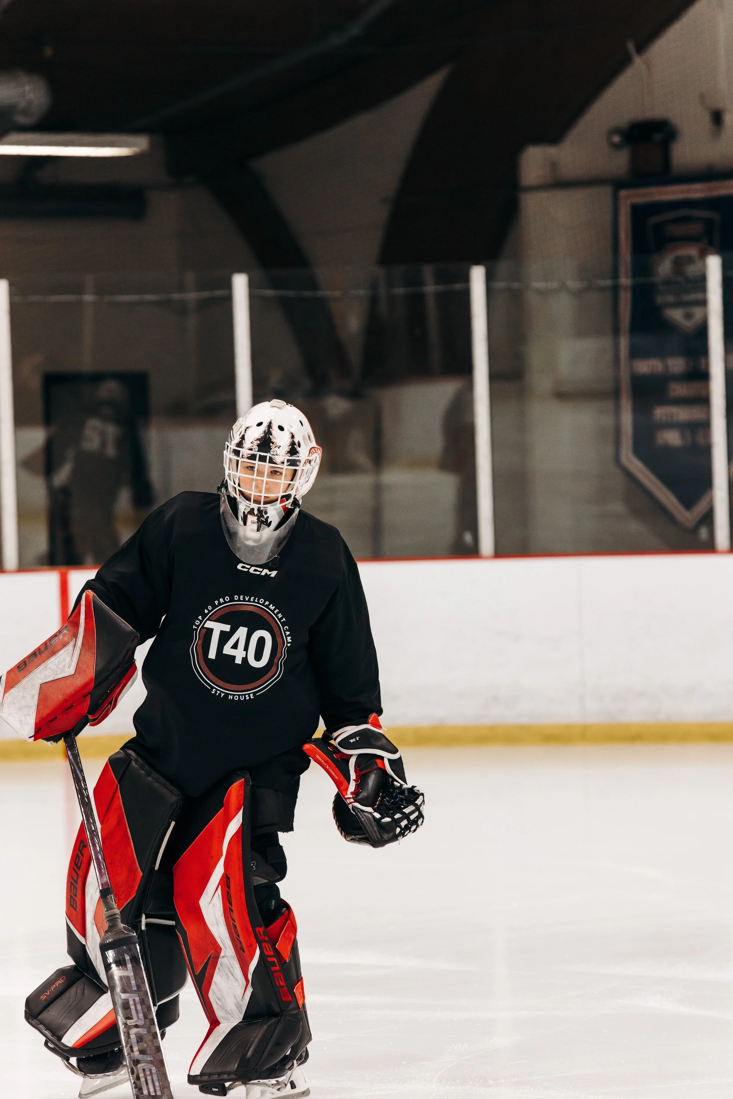 A young ice hockey goalie in black and red gear standing on the ice rink during practice. The goalie is wearing a helmet with a cage, a black jersey with a T40 logo, and holding a hockey stick. The background includes the rink's boards, glass, and a 